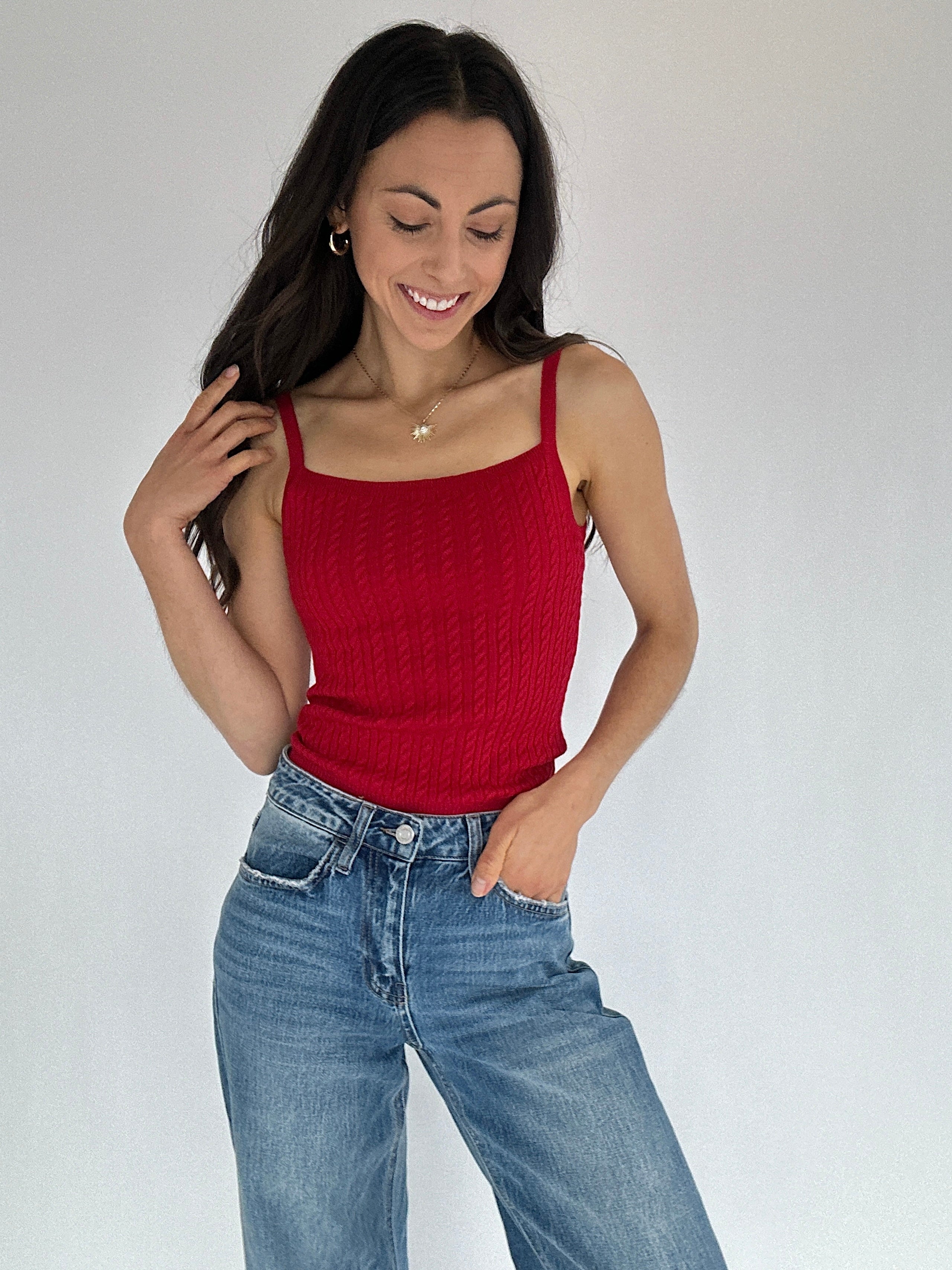 Woman wearing a red cable knit tank top and blue jeans against a plain background