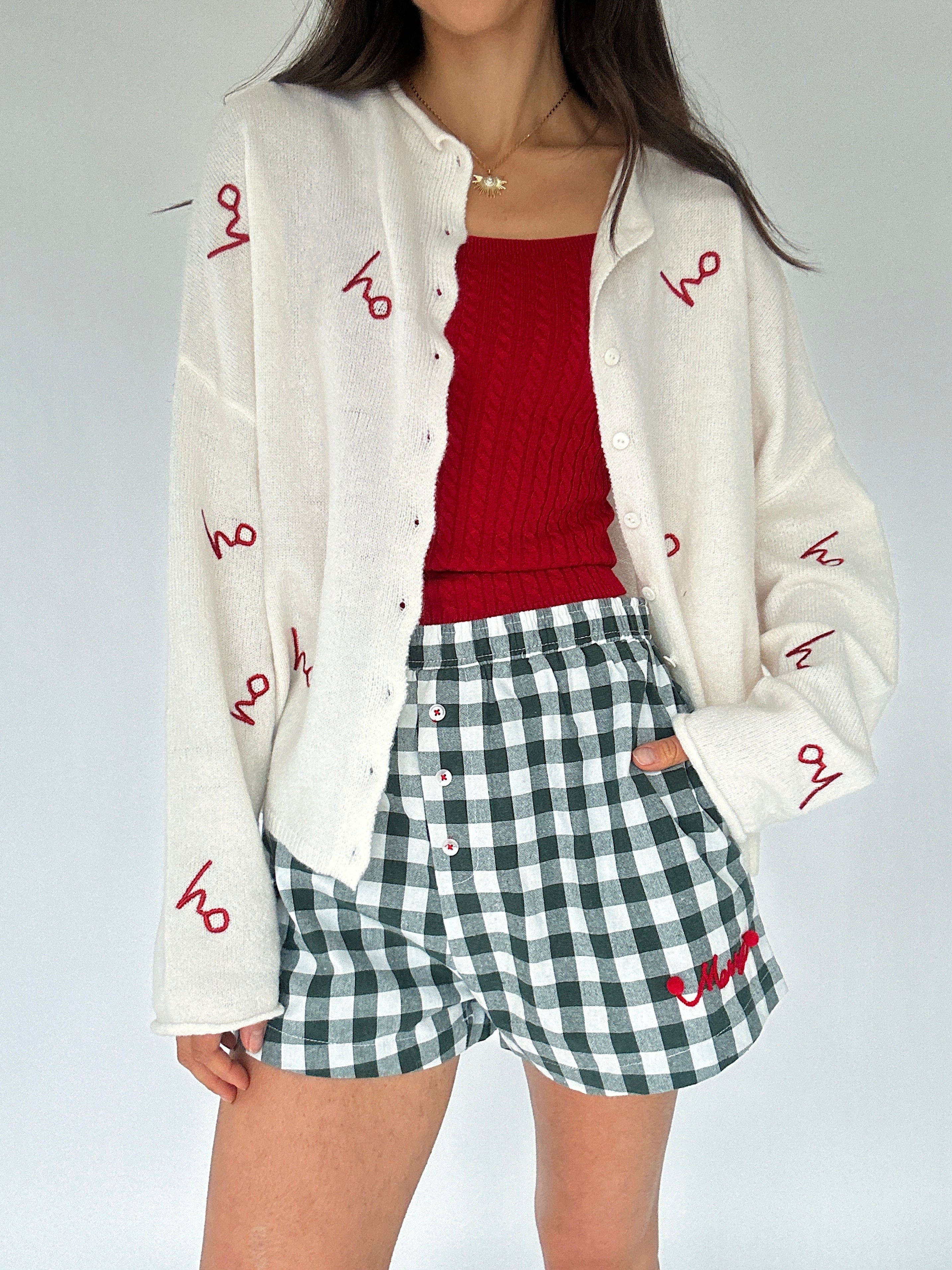 Person wearing a white cardigan with red text, red cable knit tank top, and green checkered shorts on a plain background