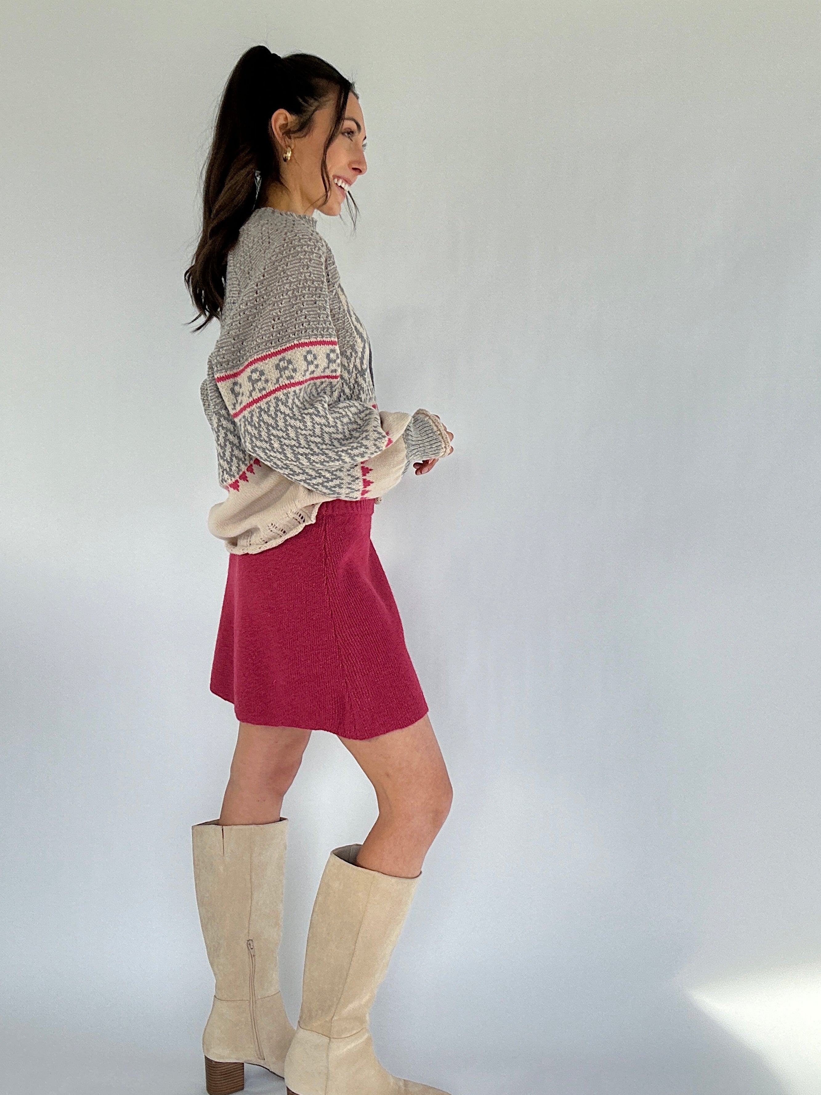 Woman wearing a patterned sweater, red skirt, and beige knee-high boots on a plain background from side
