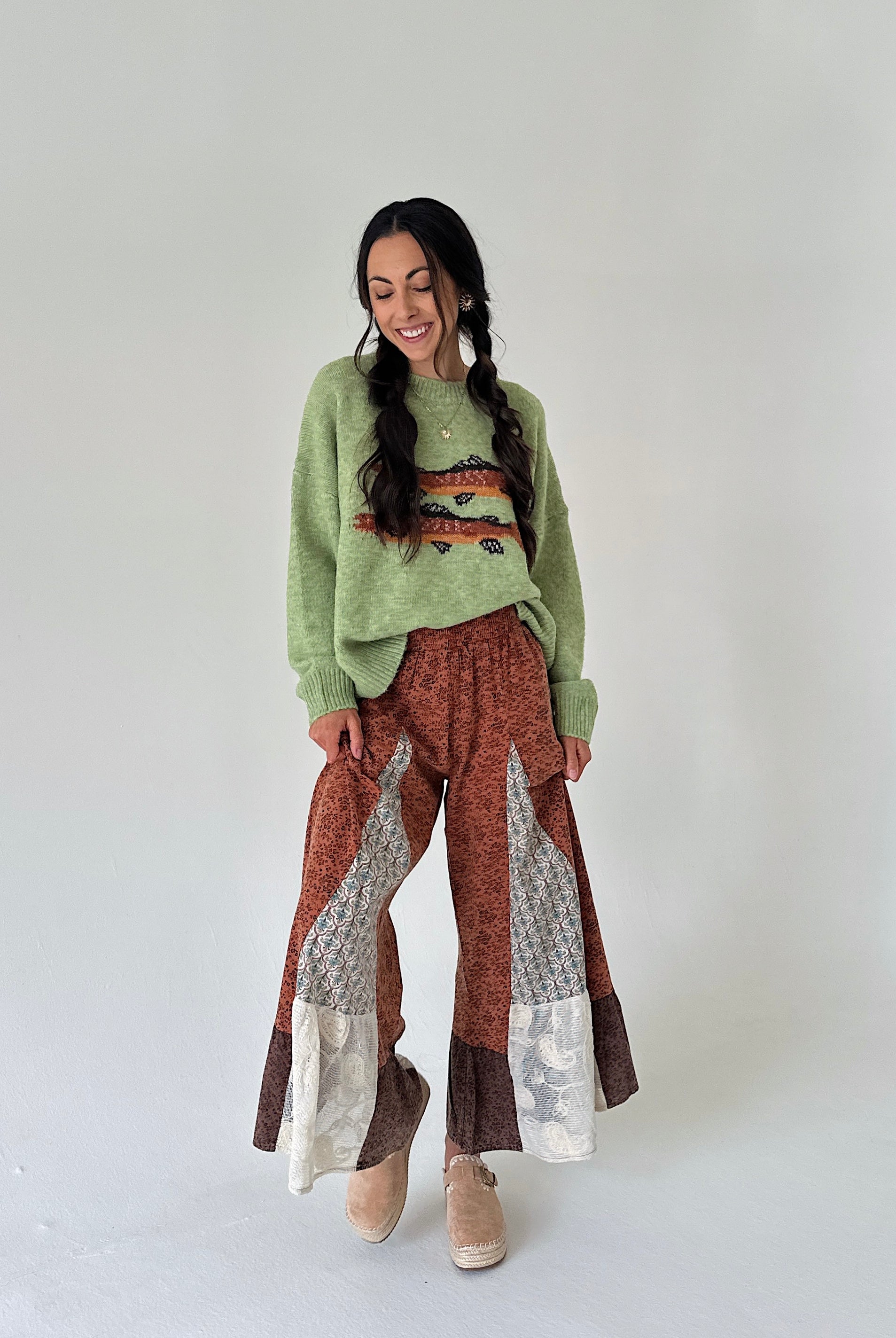 A woman modeling a light green knit sweater with a trout graphic print on the front, paired with brown and beige patterned wide-leg pants and tan clogs, against a plain light gray studio background.
