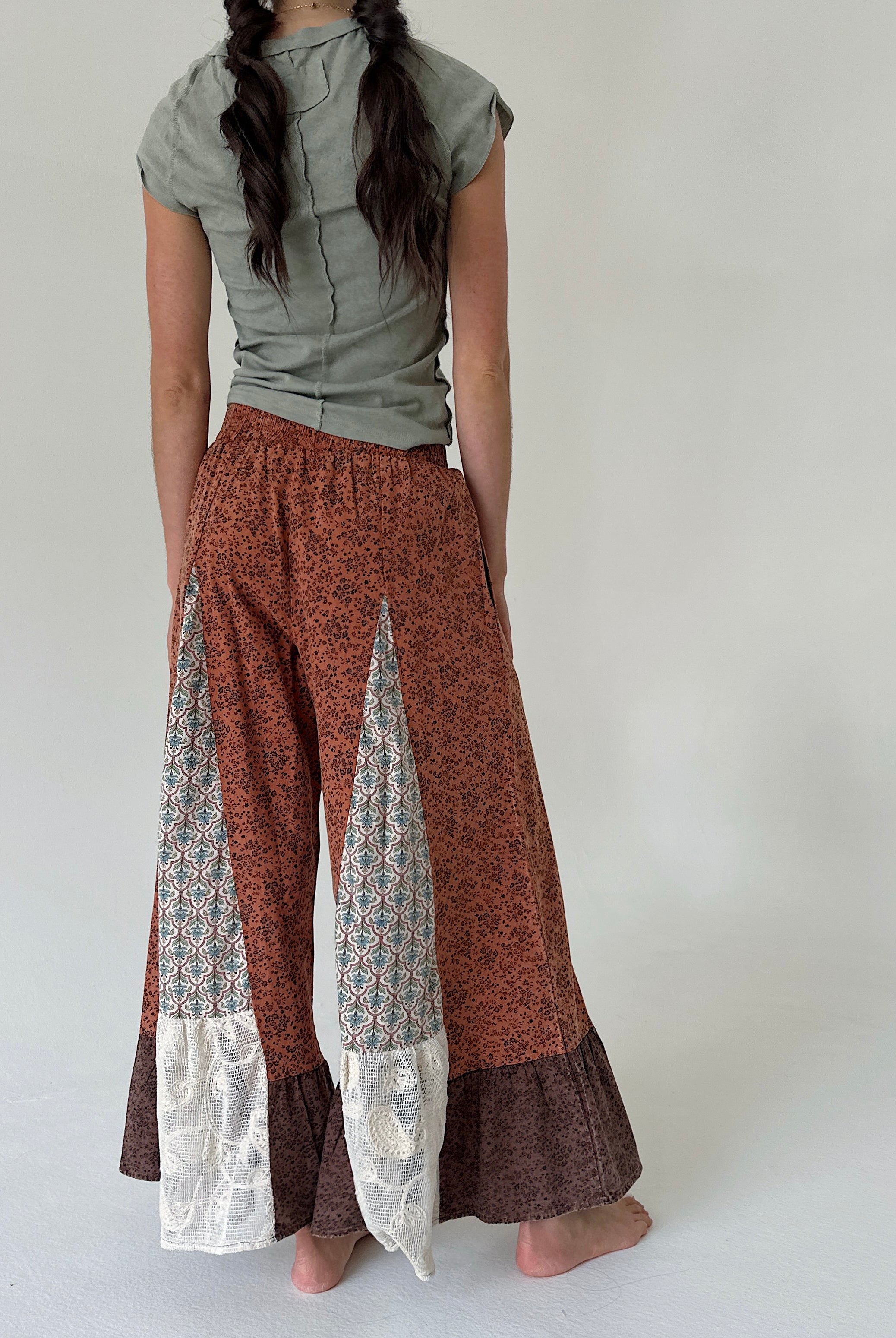 A woman shown from behind wearing wide-leg patchwork pants in rust orange, brown, and white lace, paired with an olive green top, against a plain light gray background.