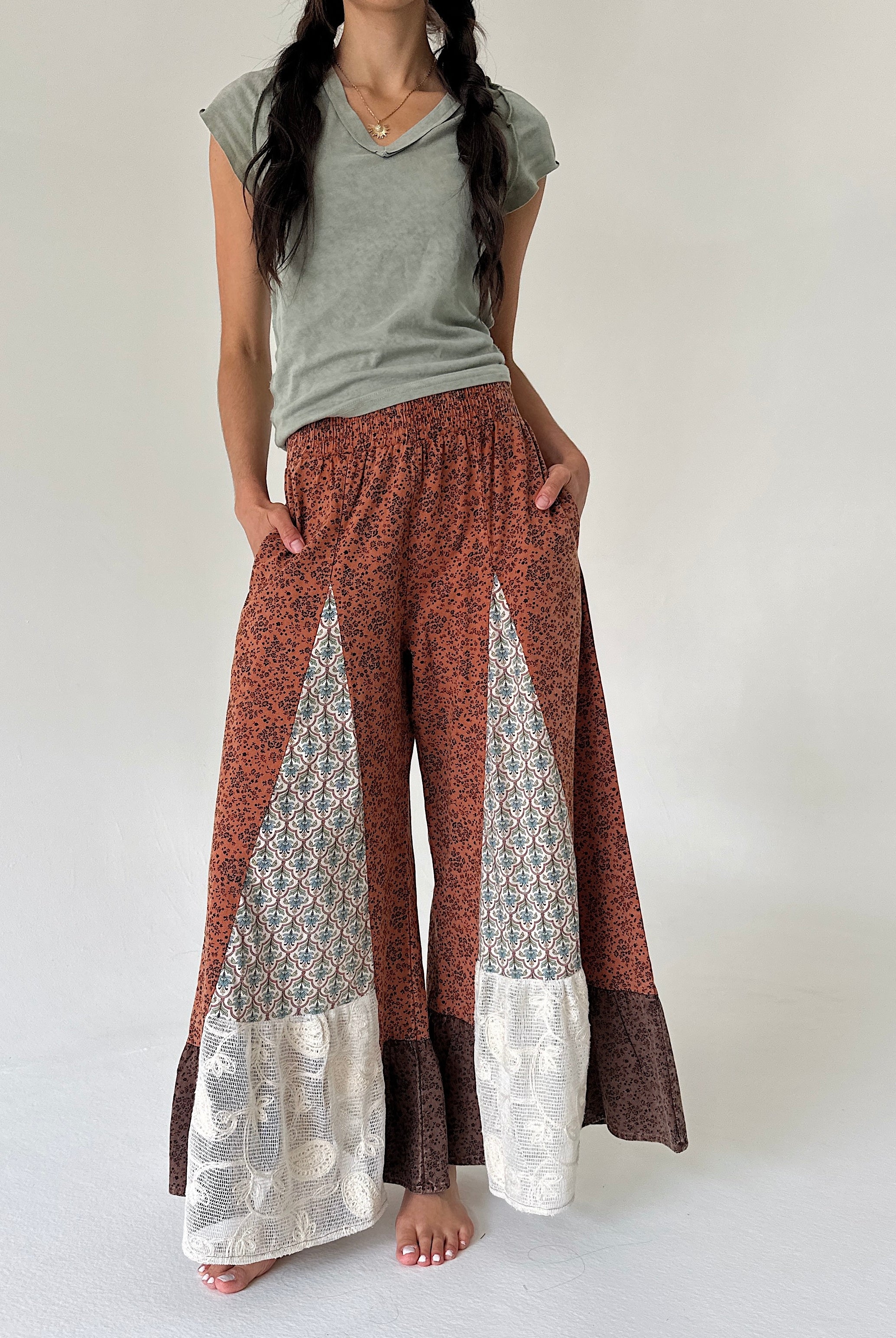 A model wearing wide-leg patchwork pants in rust red, brown, and cream, paired with a sage green t-shirt, standing against a plain off-white studio background.