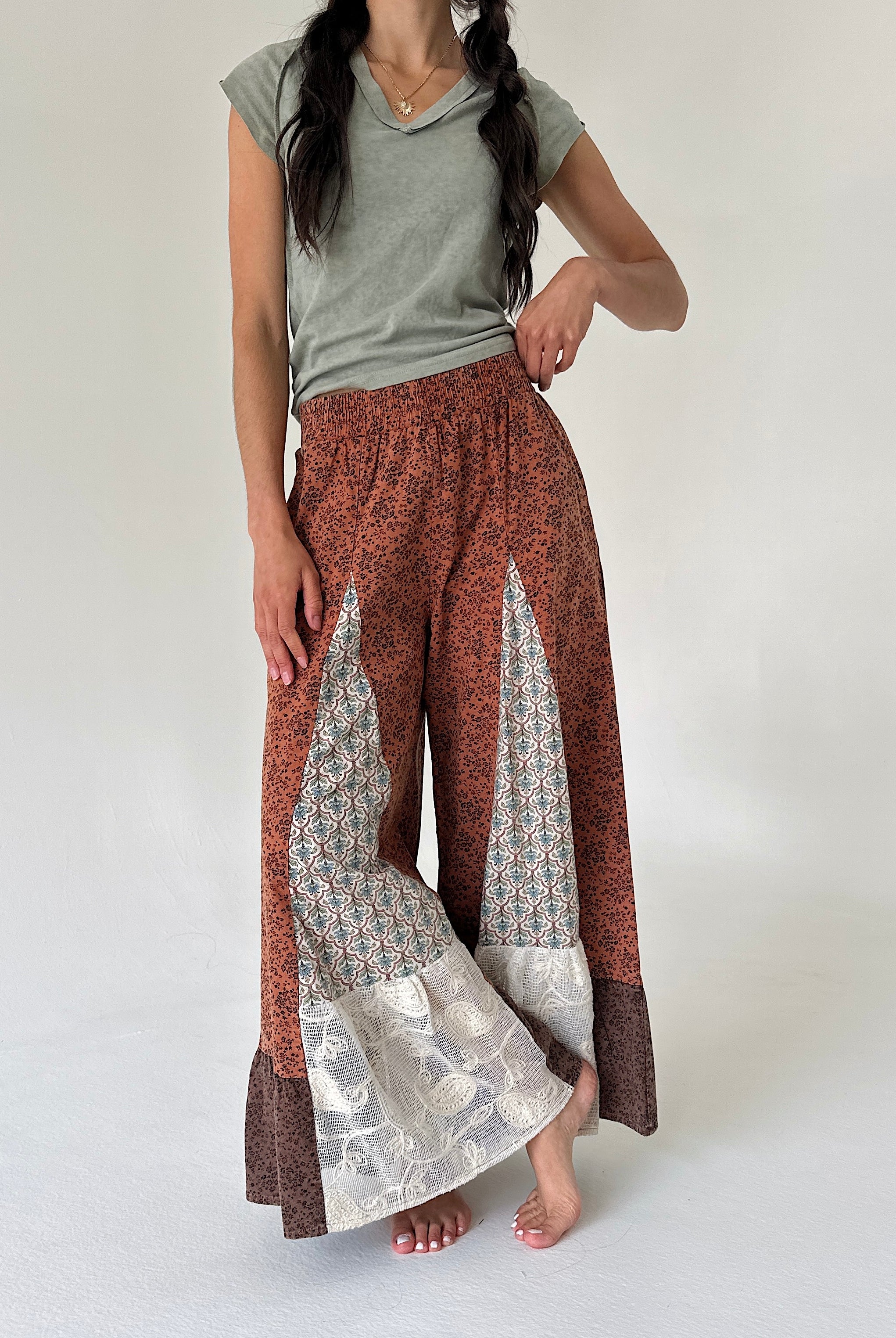 A woman modeling wide-leg patchwork pants with a rust-brown base, geometric patterned panels, lace floral accents, paired with a sage green t-shirt against a plain light gray background.