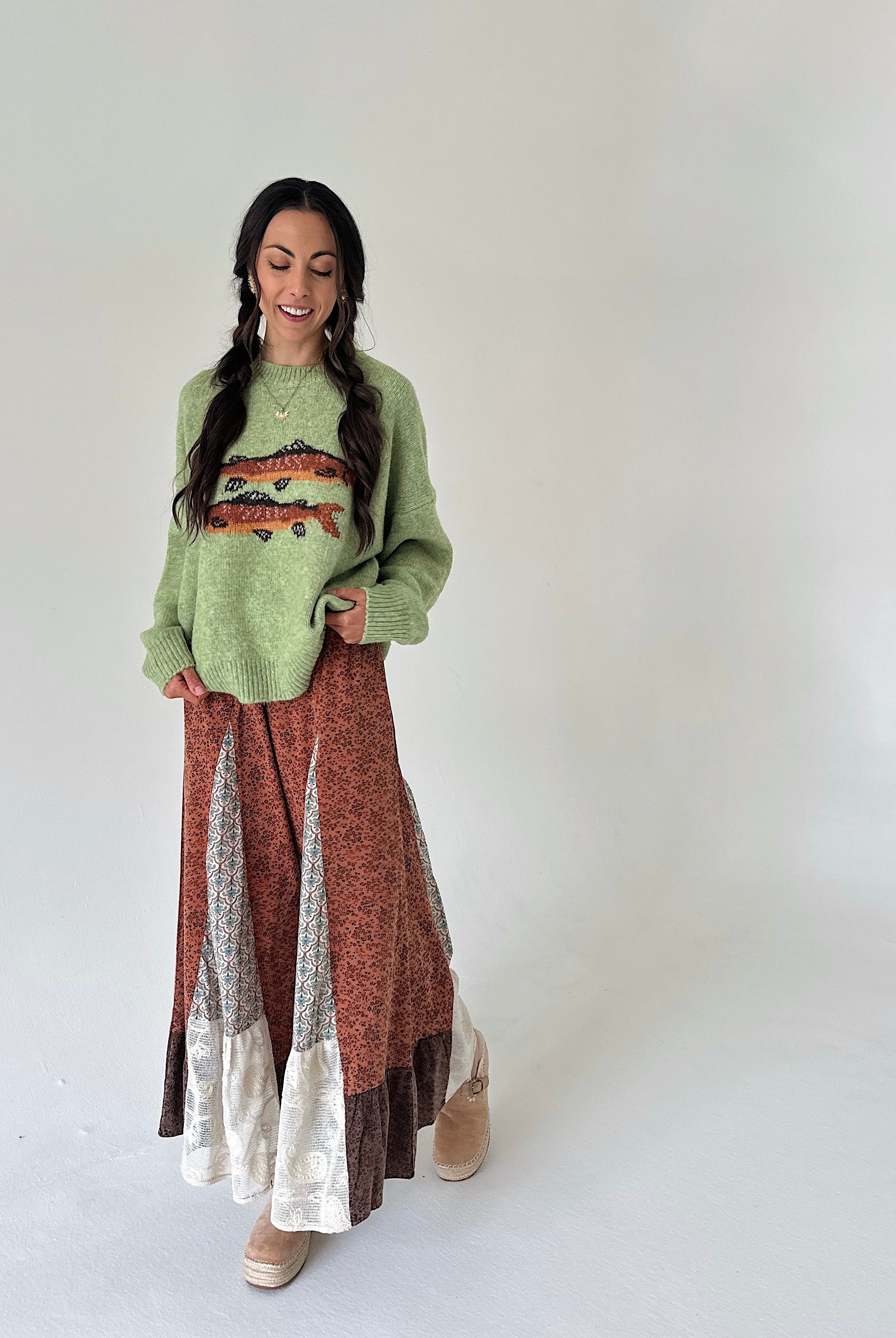 A woman modeling a cozy, long-sleeve knit sweater and a long patchwork maxi skirt. The sweater is a light sage green with a colorful fish design on the chest.