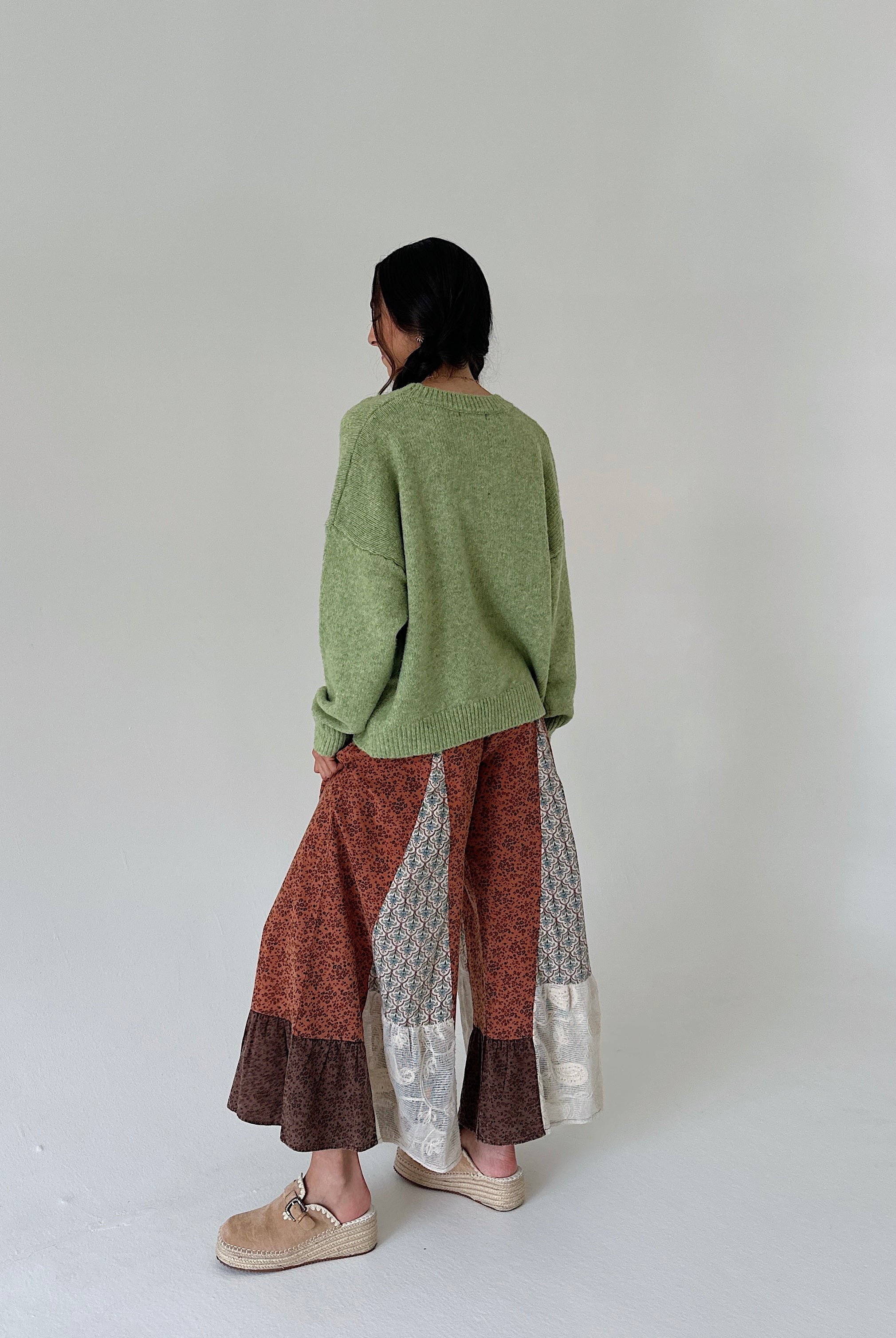 A person shown from the back wearing an oversized sage green knit sweater, wide-leg patchwork pants in rust, brown, and silver-gray, and beige slip-on shoes against a plain light gray studio background.