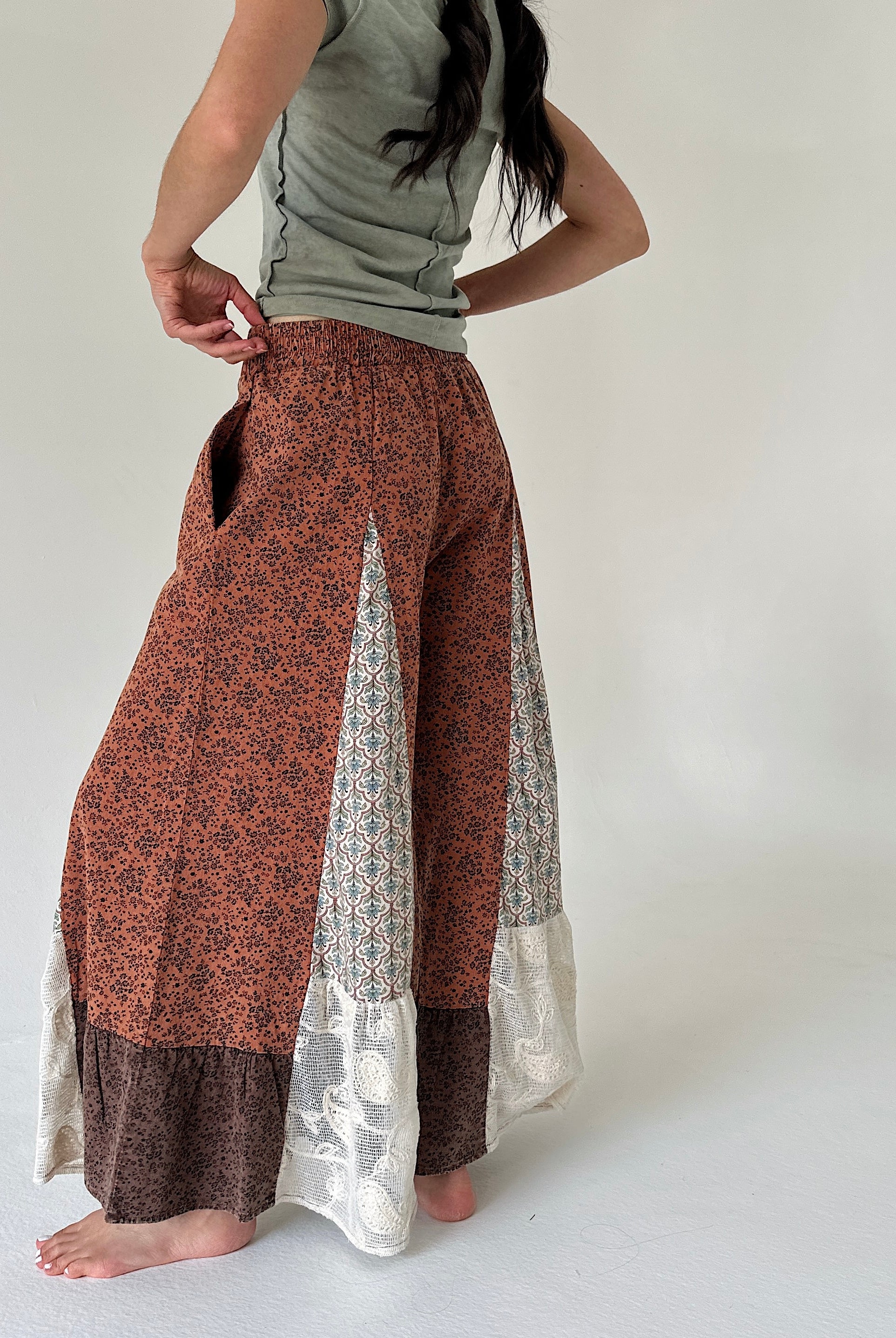 A woman wearing a pair of wide-leg patchwork pants in rust orange, brown, and cream, paired with a sage green top, shown from the back against a plain off-white background.