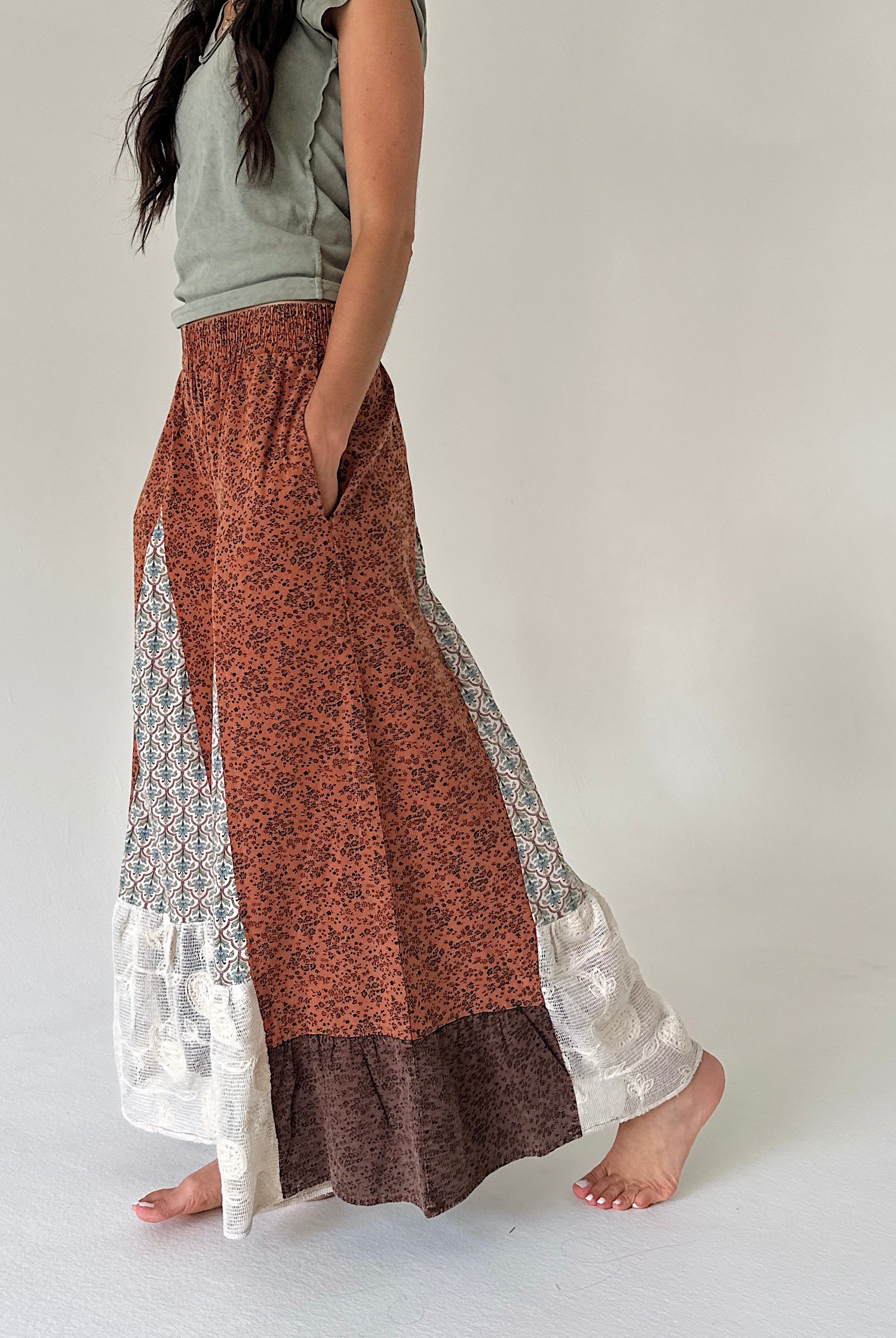A woman wearing wide leg patchwork pants with rust orange, brown, and white patterns, paired with a light gray top, standing barefoot against a plain off-white background.