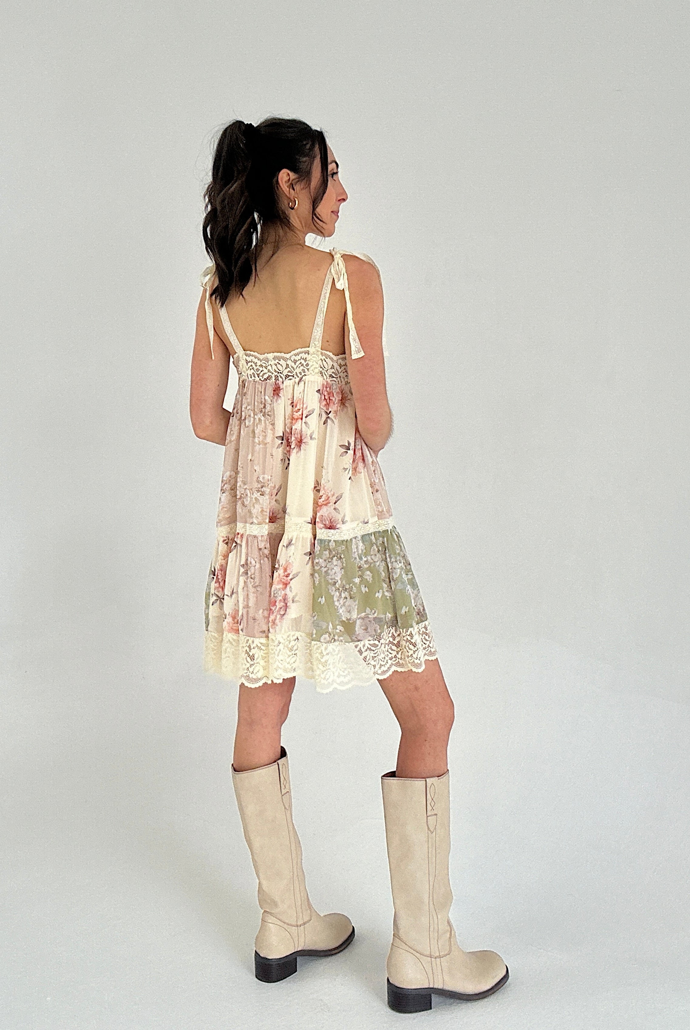 Woman wearing a floral dress with lace details and beige boots on a white background showing the back