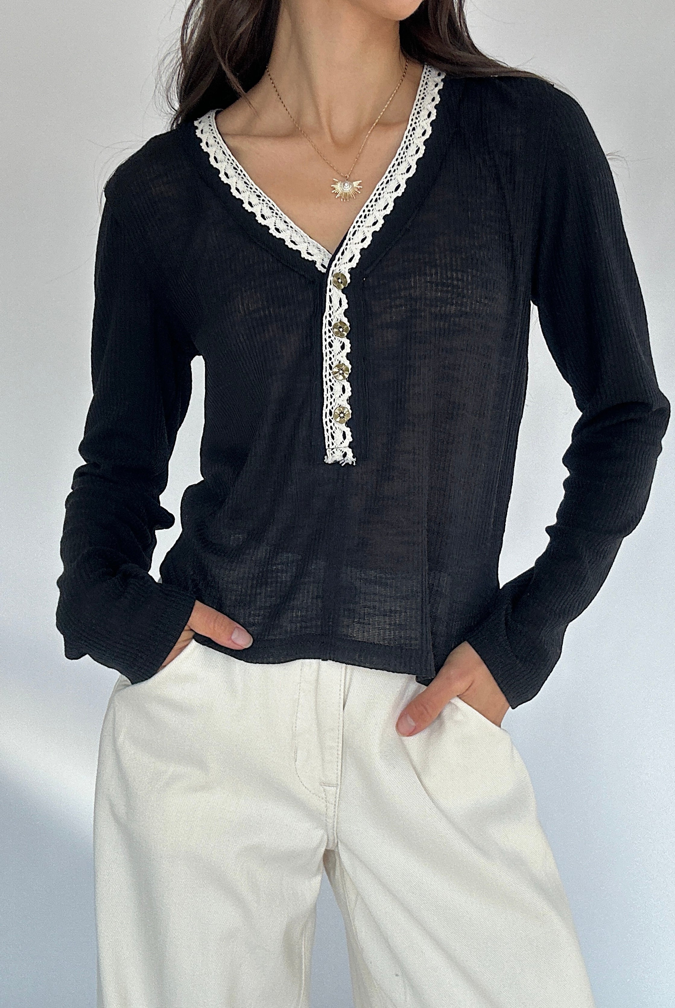 Person wearing a black long-sleeve top with white lace details and white pants on a light gray background