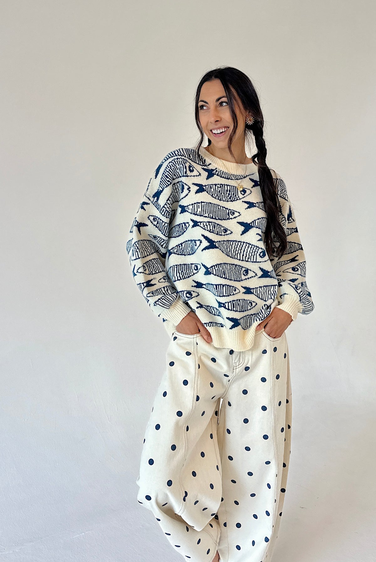 A woman modeling a cream-colored crewneck sweater with a navy blue fish pattern, paired with cream barrel-style pants with dark blue polka dots, set against a plain light gray studio background.