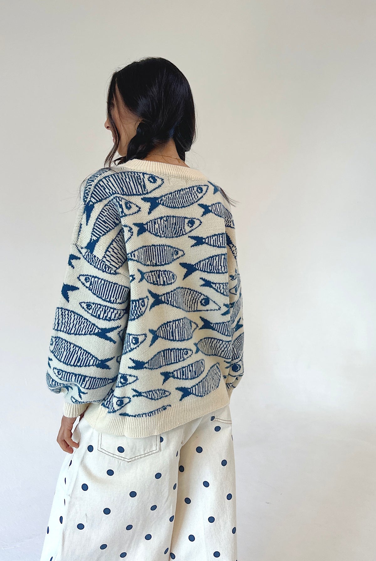 A woman shown from behind wearing a cream-colored knit sweater with a blue fish pattern and cream wide-leg pants with blue polka dots, set against a plain off-white wall.