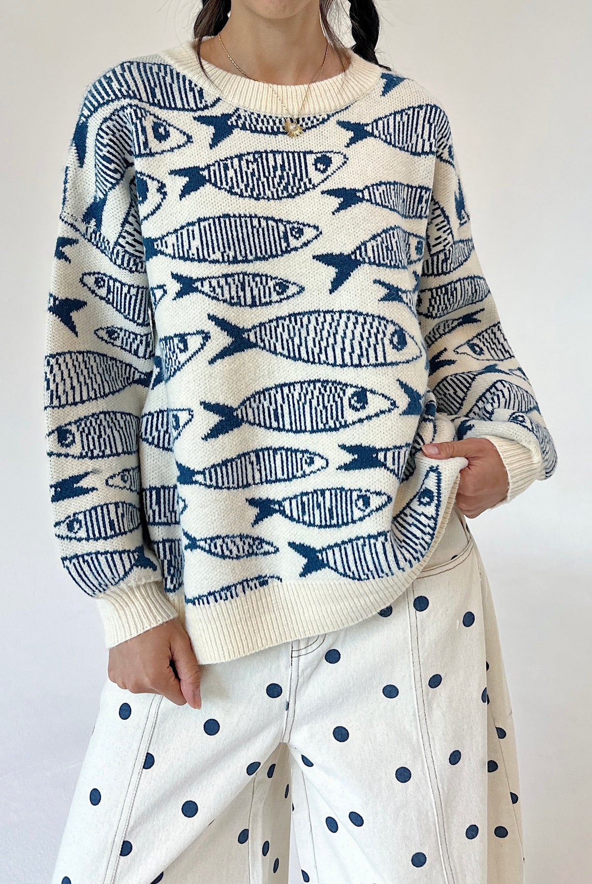A person wearing a cream knit sweater with a repeating blue fish pattern, paired with cream pants featuring dark blue polka dots, against a plain light background.