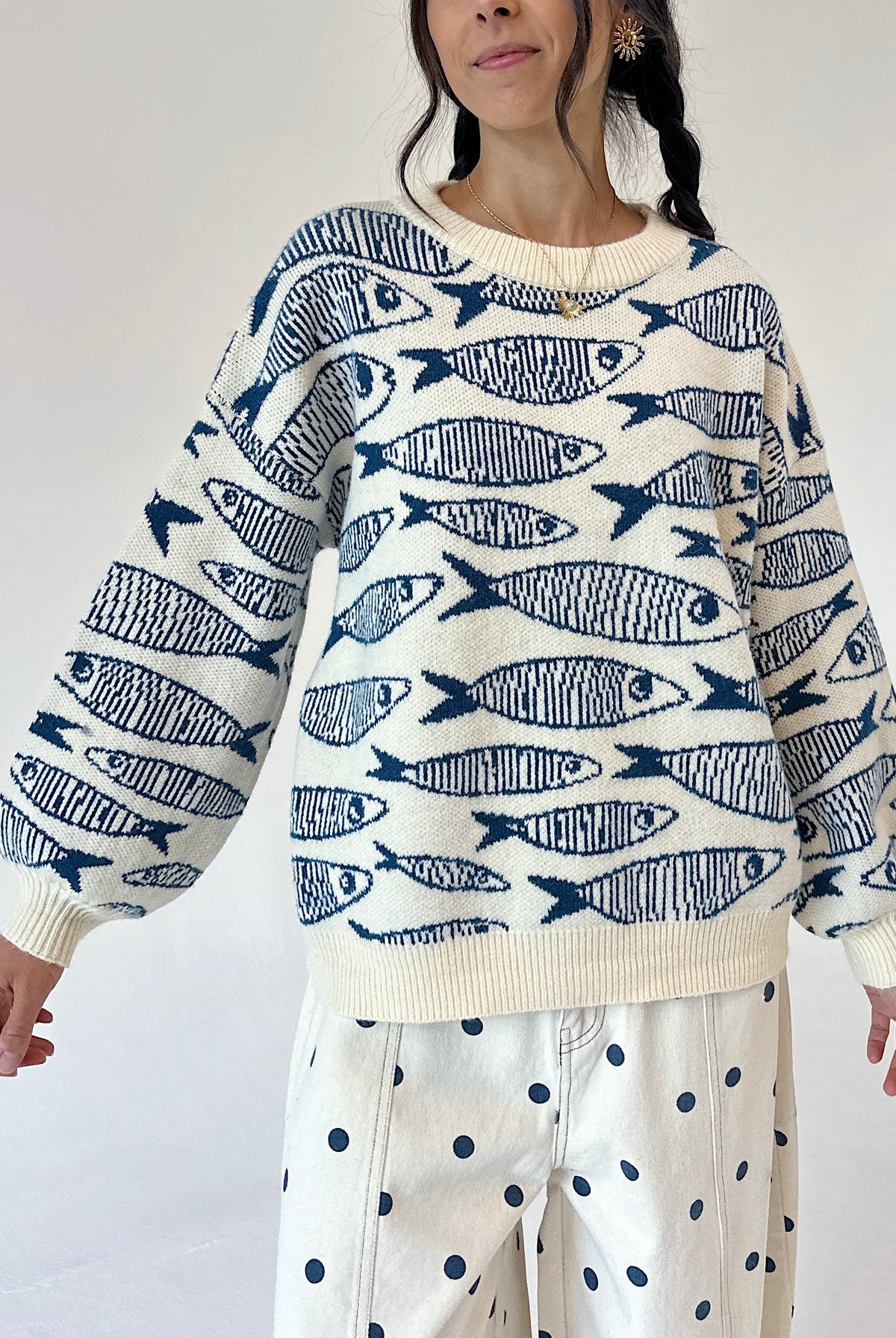 A woman wearing a cream-colored knit sweater with a navy blue fish pattern, paired with cream pants featuring navy blue polka dots, against a plain white background.