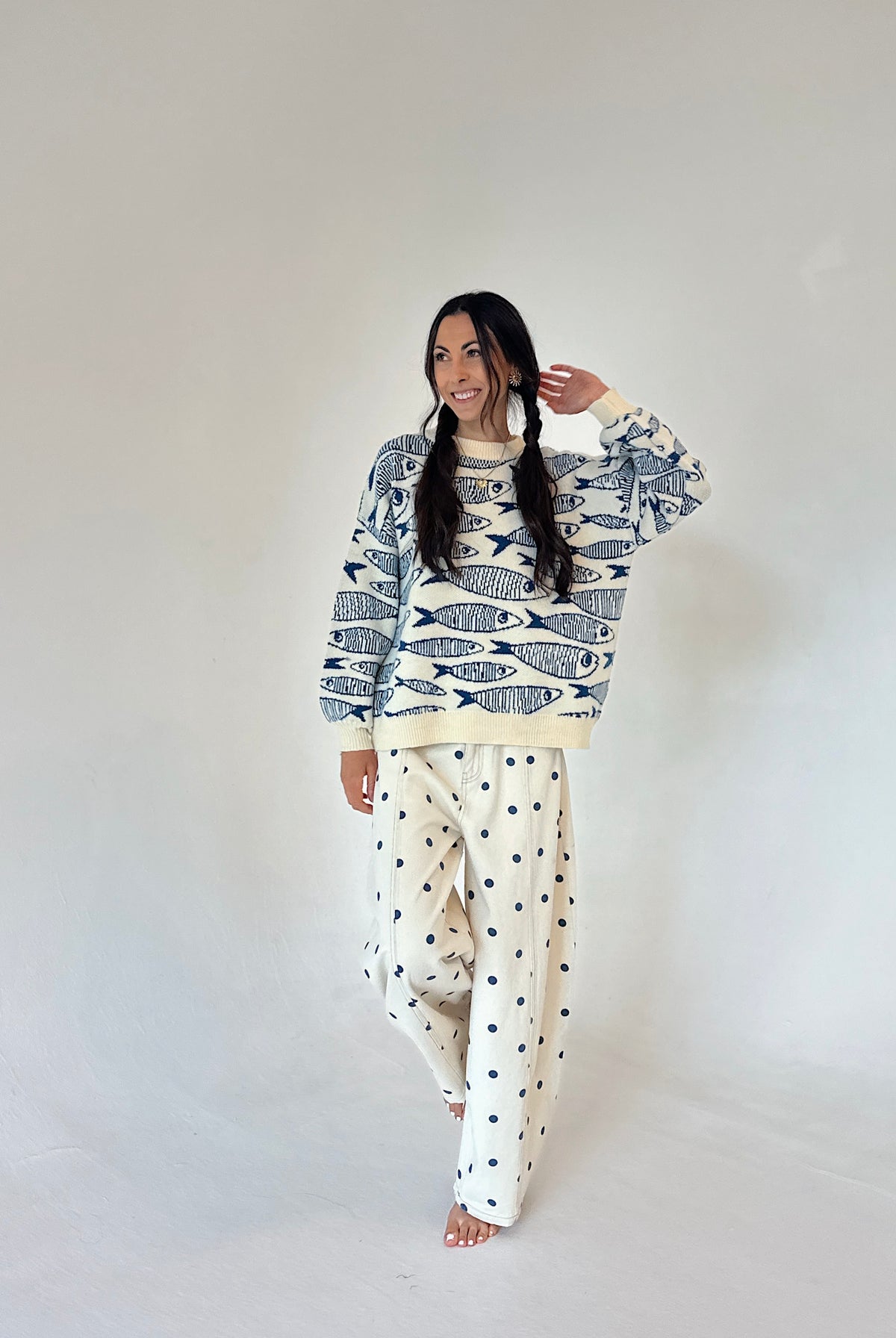 A woman modeling a long-sleeve crewneck sweater with a navy blue fish pattern on an off-white background, and off-white pants with a navy blue polka dot pattern, set against a plain light gray studio background.