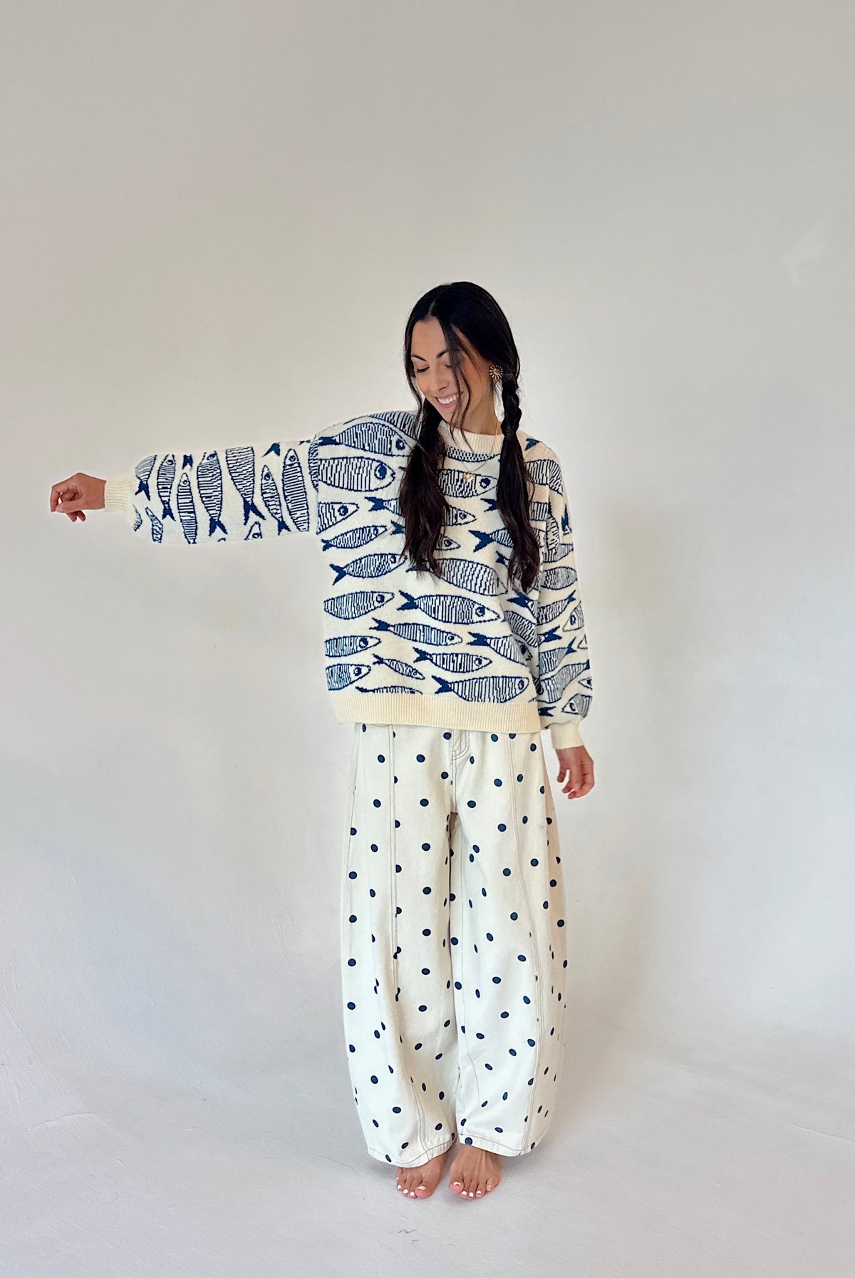 A woman modeling a long-sleeve sweater with a blue and white fish pattern and wide-leg pants with a small blue polka dot pattern on an off-white background, set against a plain light gray studio backdrop.