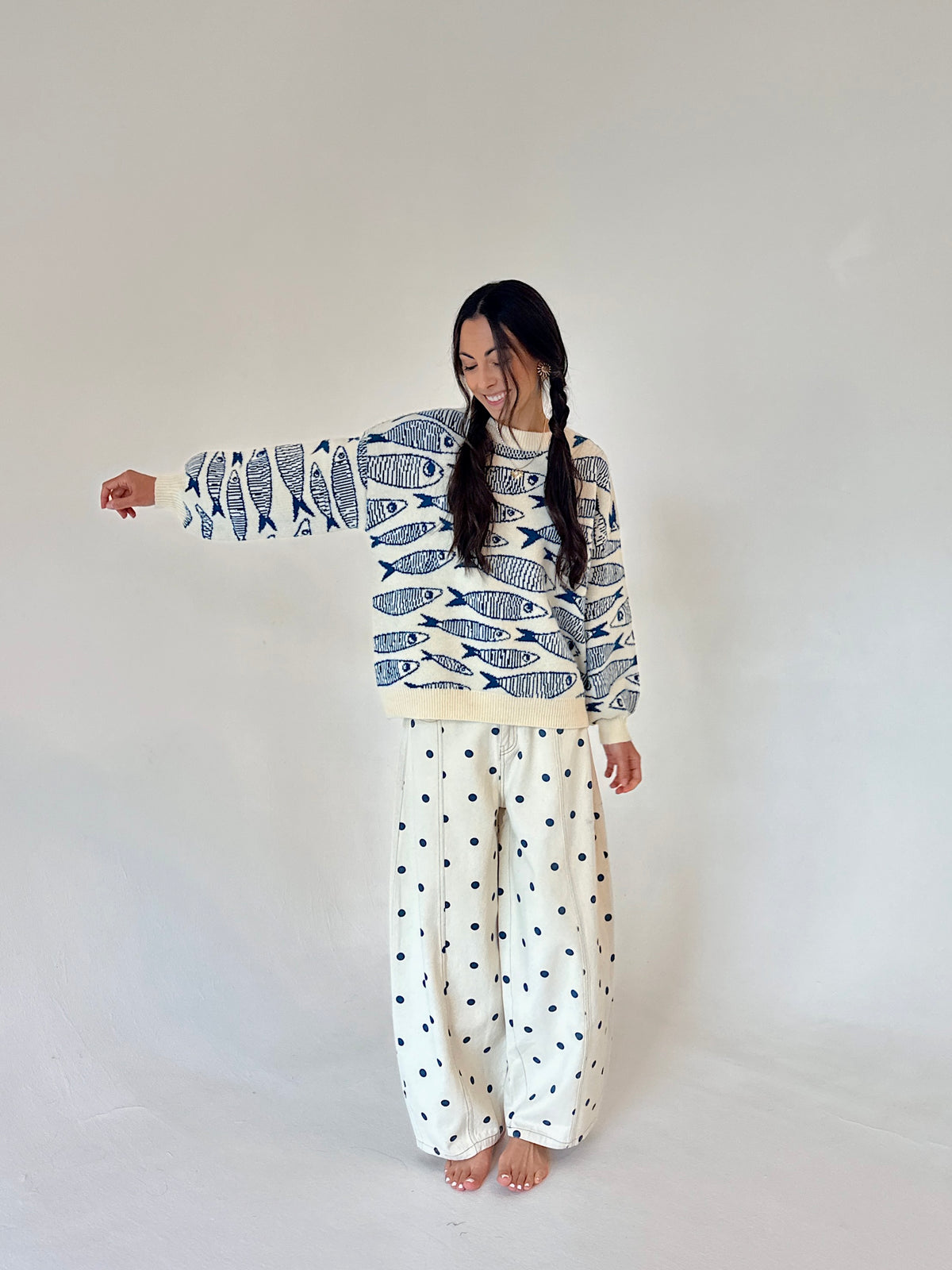 A woman modeling a long-sleeve sweater with a blue and white fish pattern and wide-leg pants with a small blue polka dot pattern on an off-white background, set against a plain light gray studio backdrop.