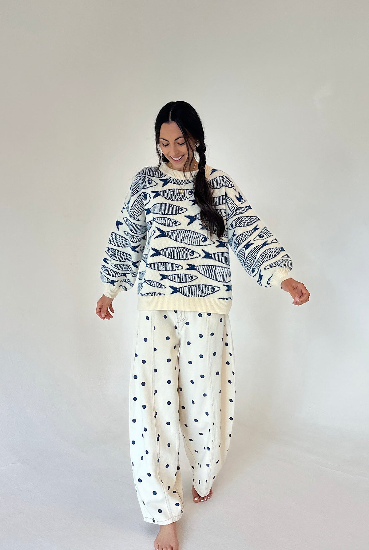 A woman modeling a cream sweater with a navy fish pattern and cream pants with navy polka dots, set against a plain light gray studio background.