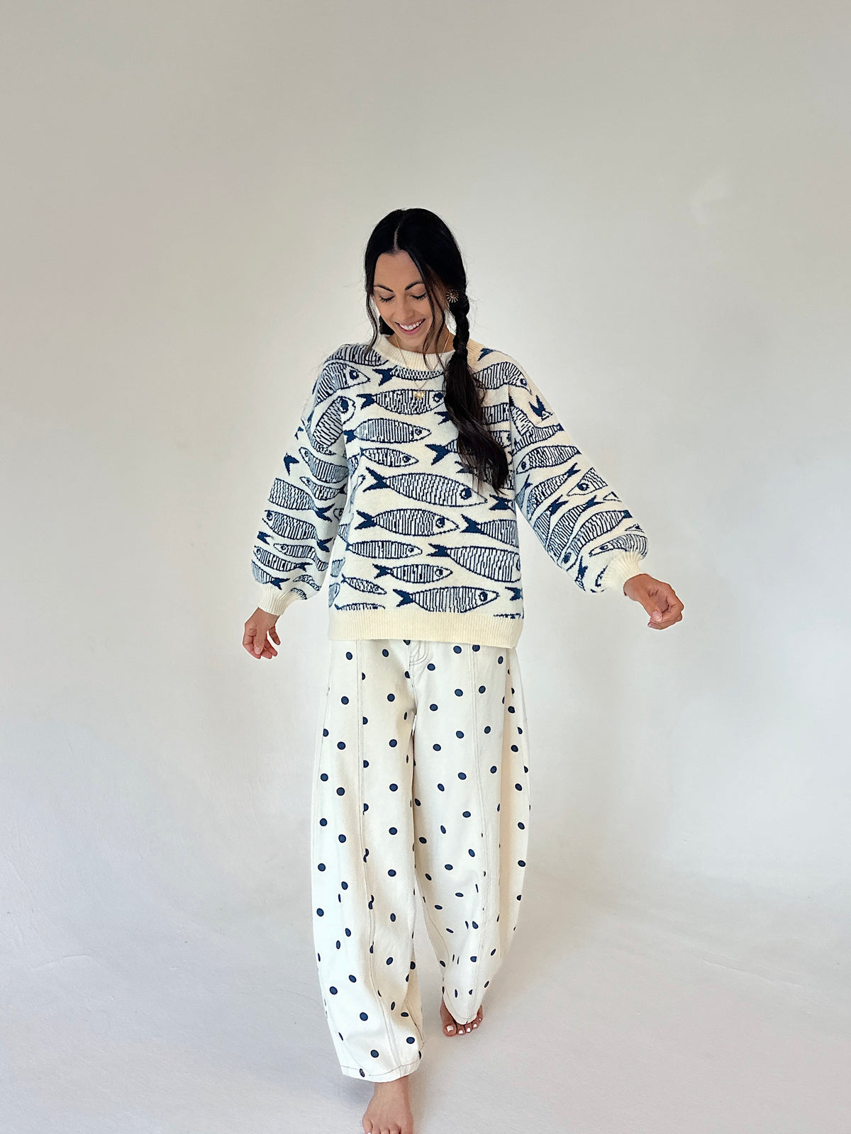 A woman modeling a cream sweater with a navy fish pattern and cream pants with navy polka dots, set against a plain light gray studio background.