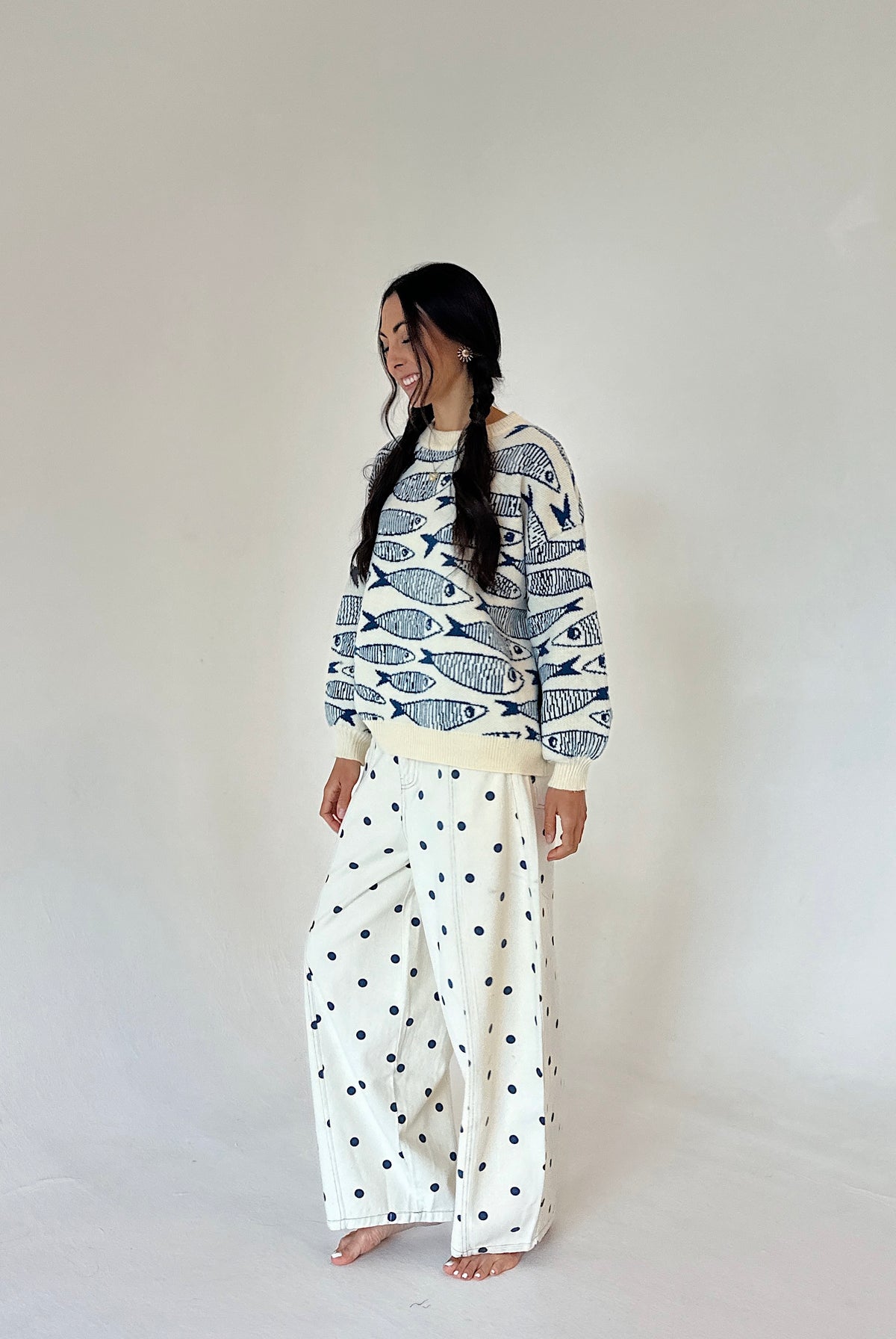 A woman modeling long-sleeve crewneck sweater with a navy blue fish pattern on an off-white background, and wide-leg pants with a navy blue polka dot pattern on an off-white background, set against a plain light gray studio backdrop.