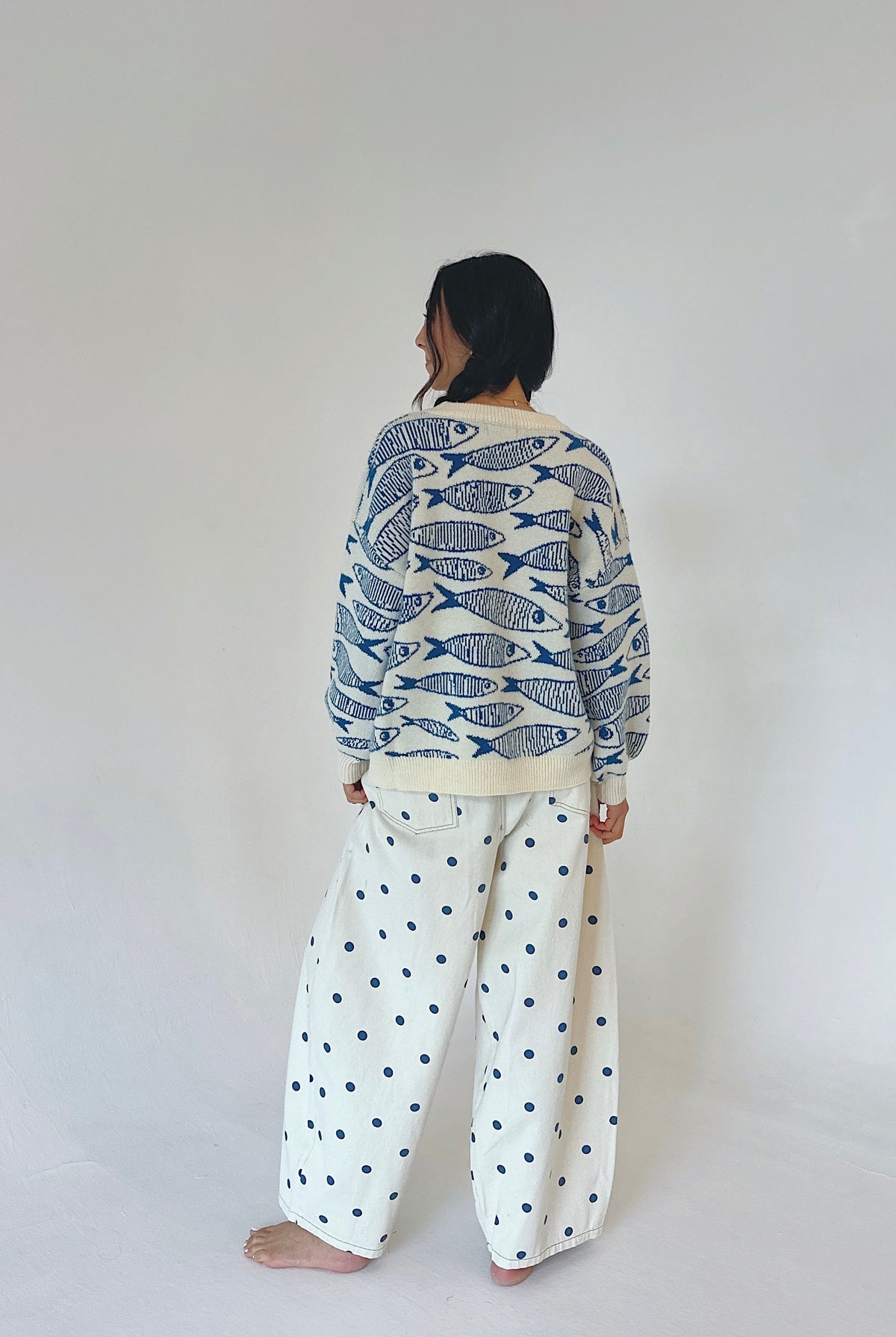 A person standing barefoot against a plain light gray wall, viewed from the back. They are wearing a cream knit sweater covered in a blue fish pattern and matching cream wide-leg pants with blue polka dots.