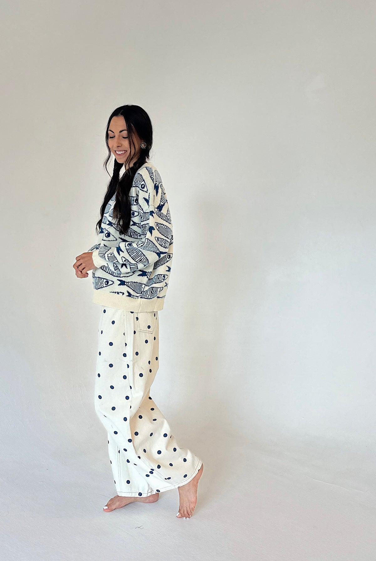 A woman stands barefoot against a plain off-white studio background, wearing a cream and navy fish patterned sweater with cream pants featuring navy polka dots.