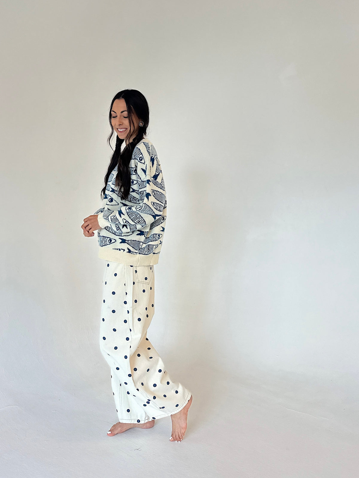A woman stands barefoot against a plain off-white studio background, wearing a cream and navy fish patterned sweater with cream pants featuring navy polka dots.