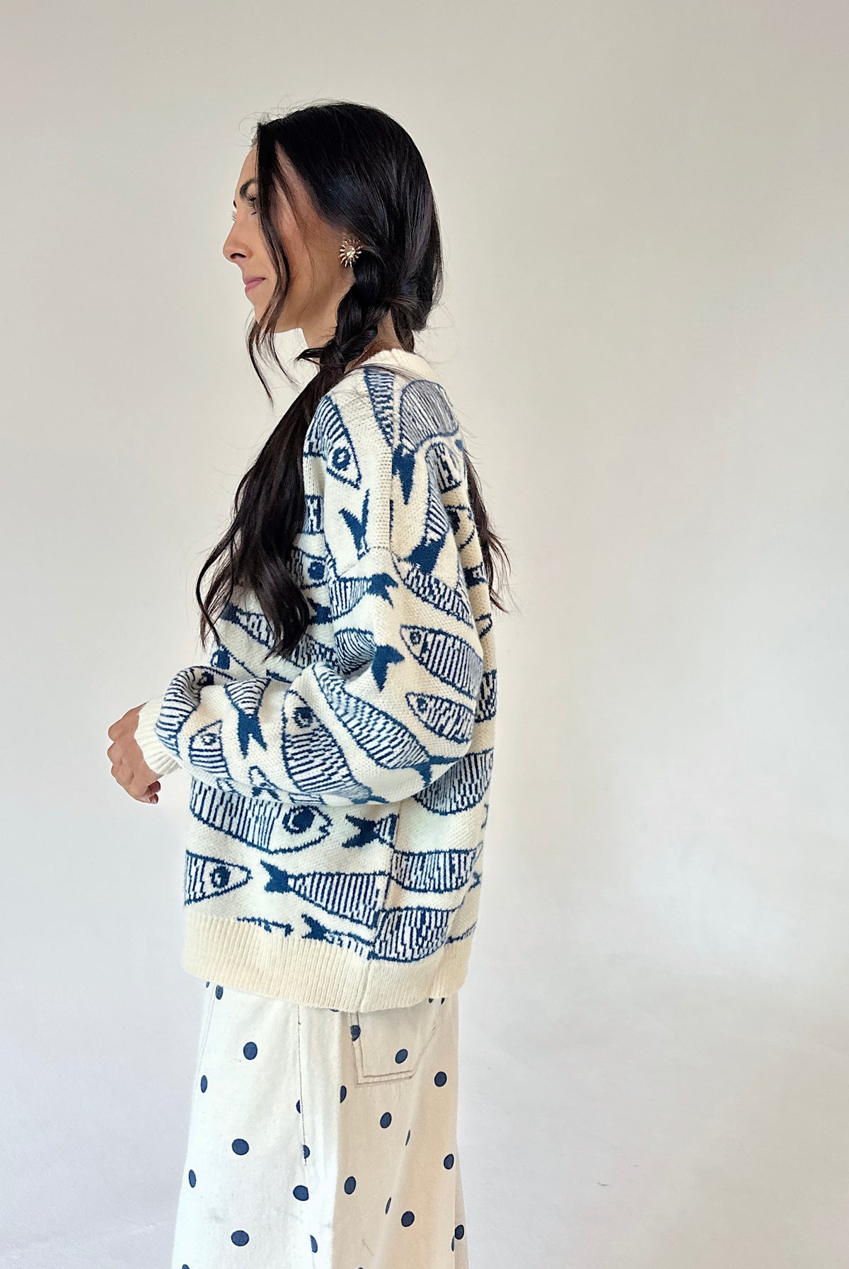 A woman shown in profile wearing a cream knit sweater with a blue fish pattern and matching cream pants with blue polka dots, standing against a plain off-white wall.