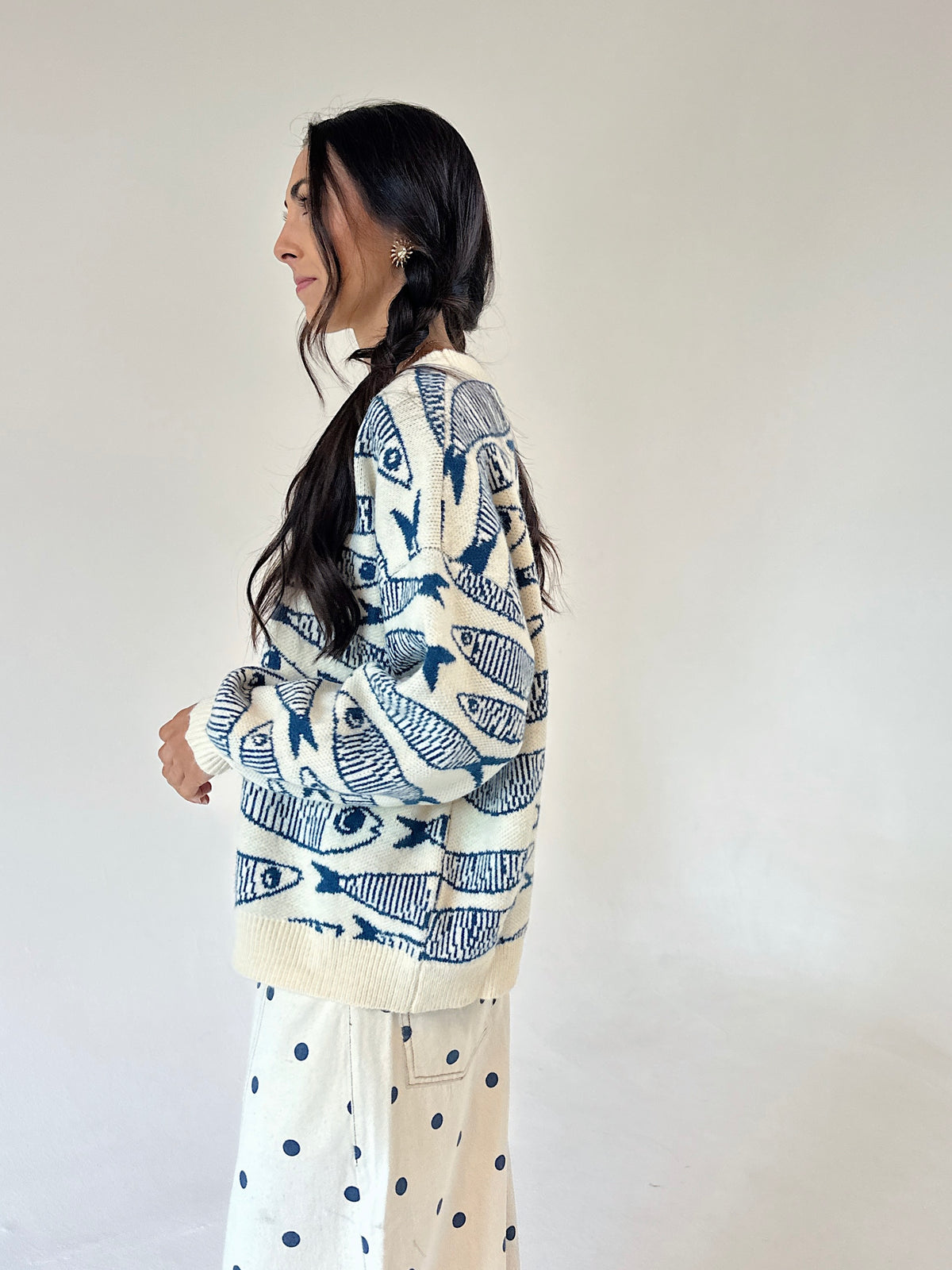 A woman shown in profile wearing a cream knit sweater with a blue fish pattern and matching cream pants with blue polka dots, standing against a plain off-white wall.