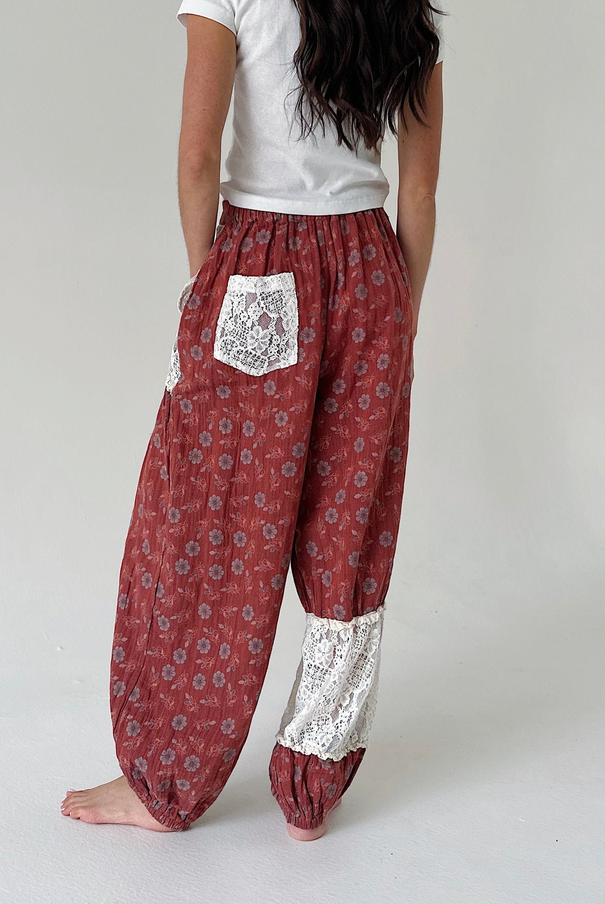 A person shown from the back wearing loose, patterned harem-style pants in a muted red-brown tone with white lace pocket and lower-leg panels, paired with a white top against a plain light gray studio background.
