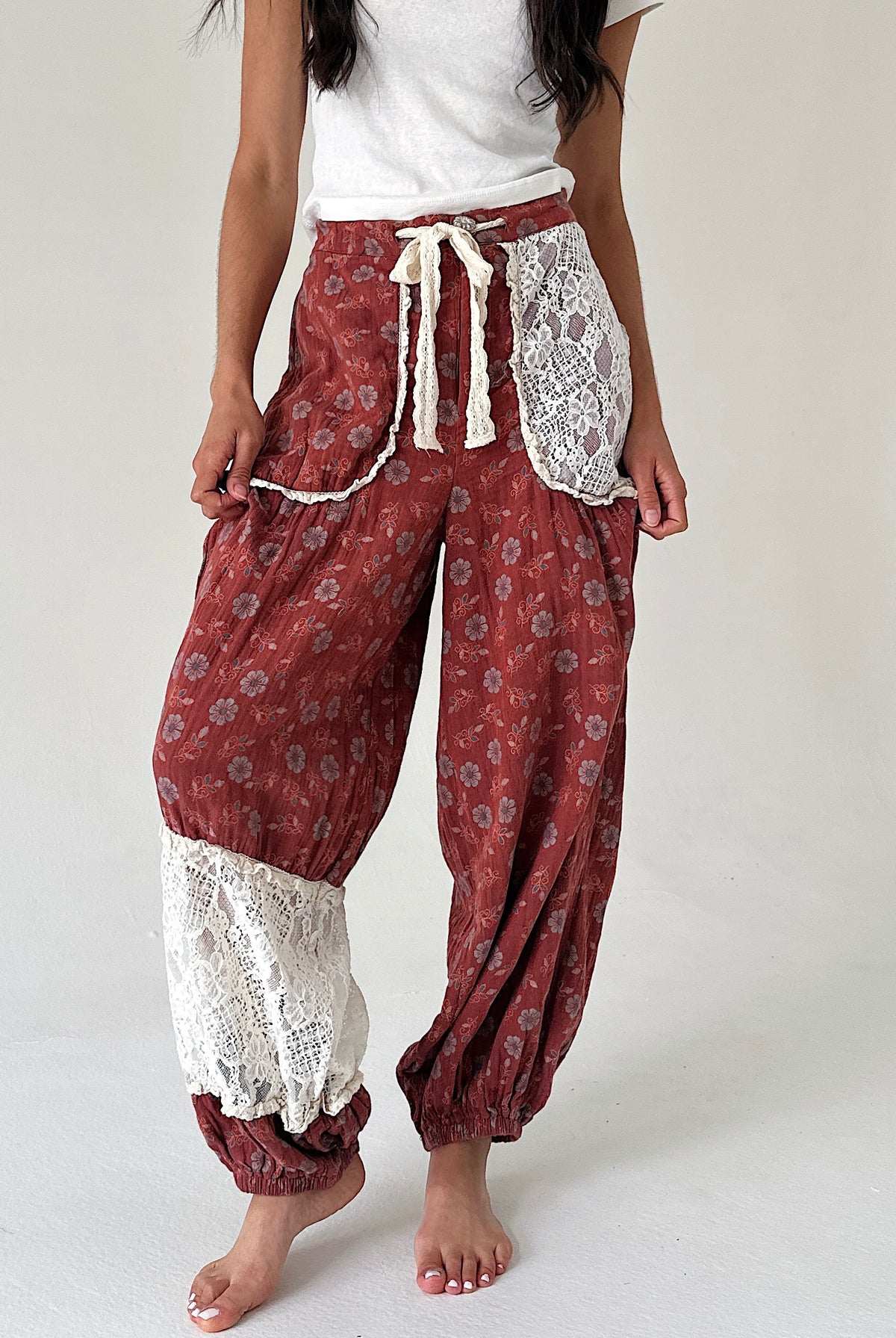 A person wearing loose, draped harem-style pants in a rust red floral print with white lace trim and a cream drawstring, paired with a white top, photographed against a plain light background.