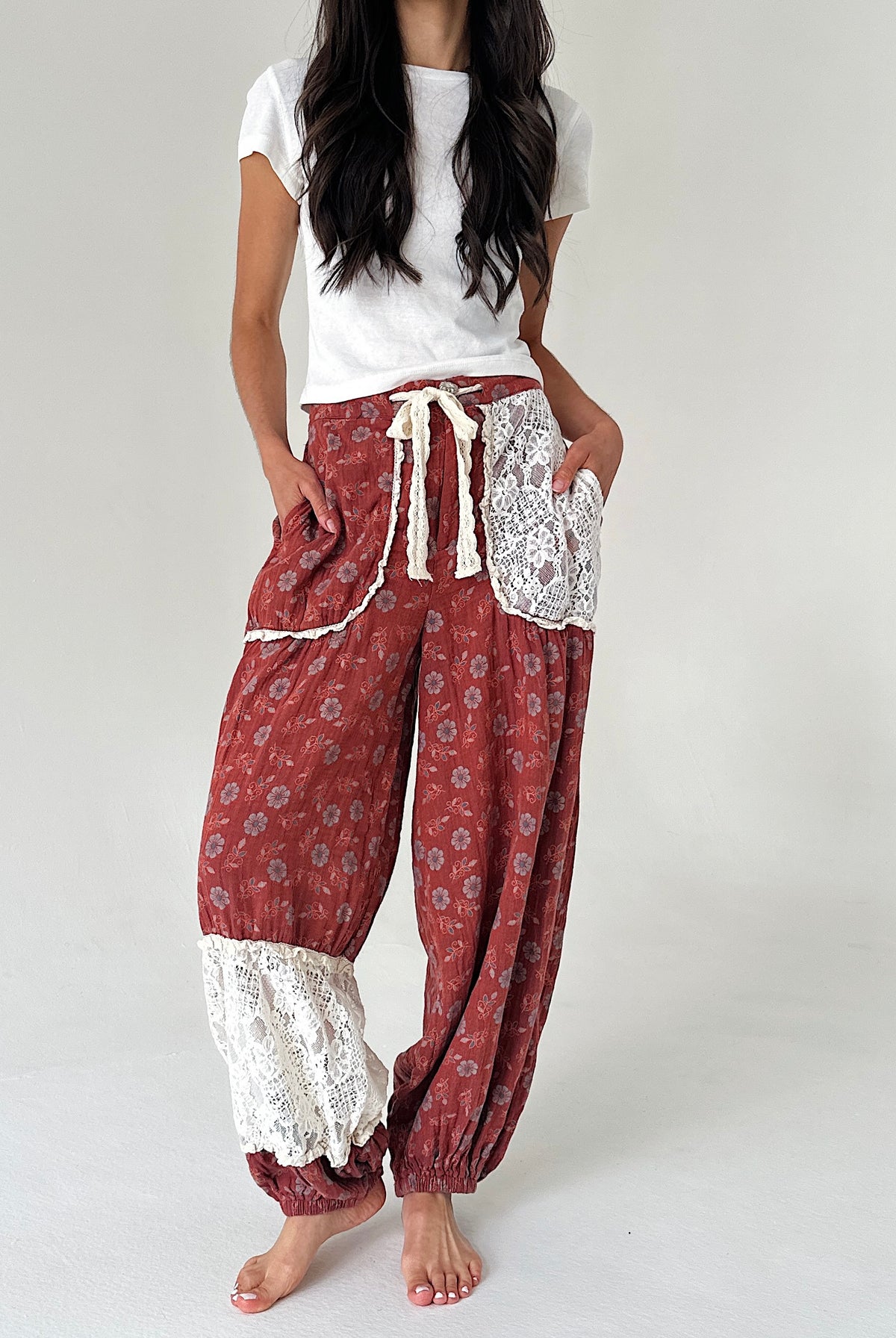 A model wearing loose, bohemian-style harem pants in a rust red floral print with white lace trim and a cream drawstring, paired with a white t-shirt, against a plain light gray background.