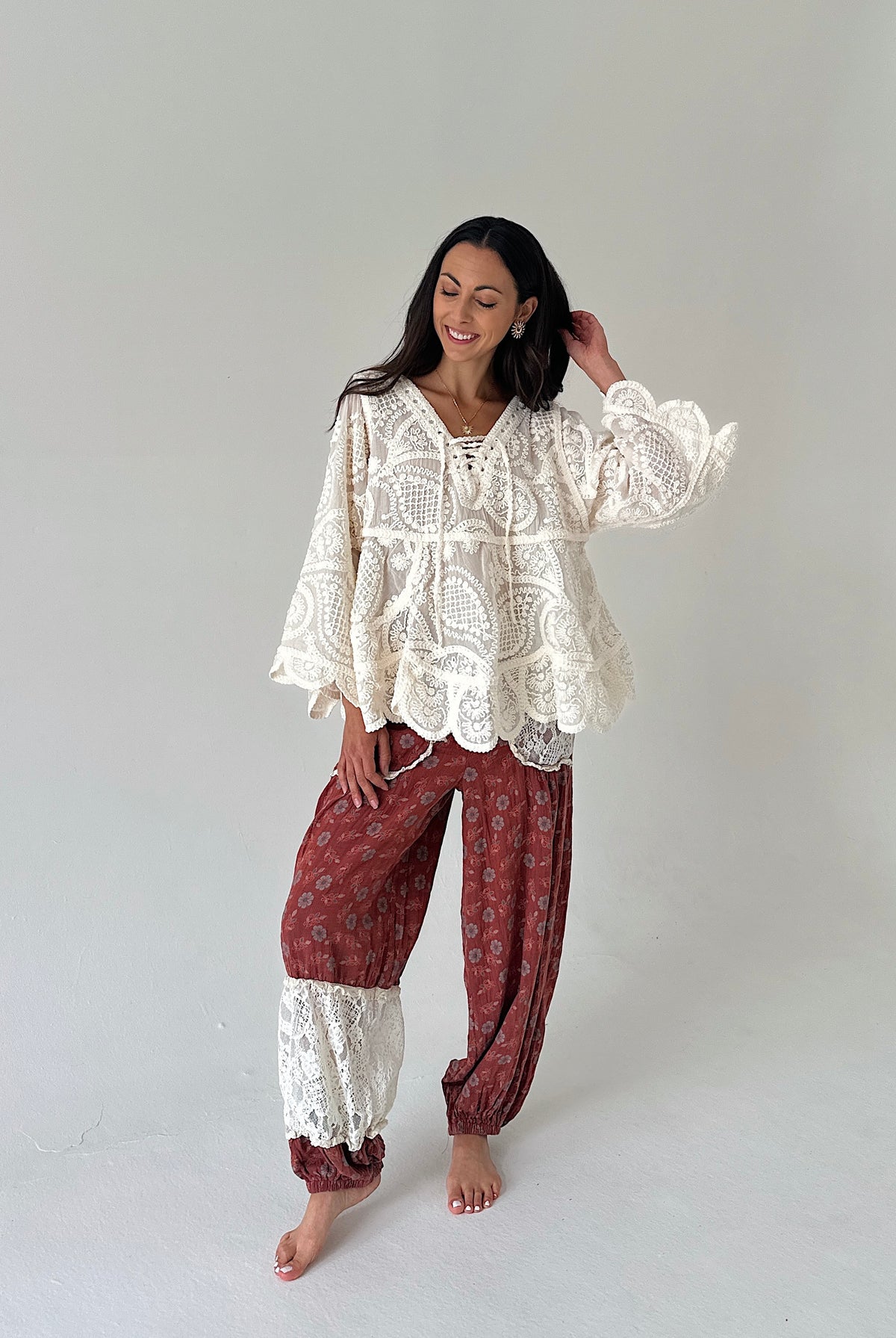 A woman modeling a bohemian-style two-piece outfit consisting of a cream lace long-sleeve top and rust-red patterned harem pants with lace cuffs, standing against a plain light gray studio background.
