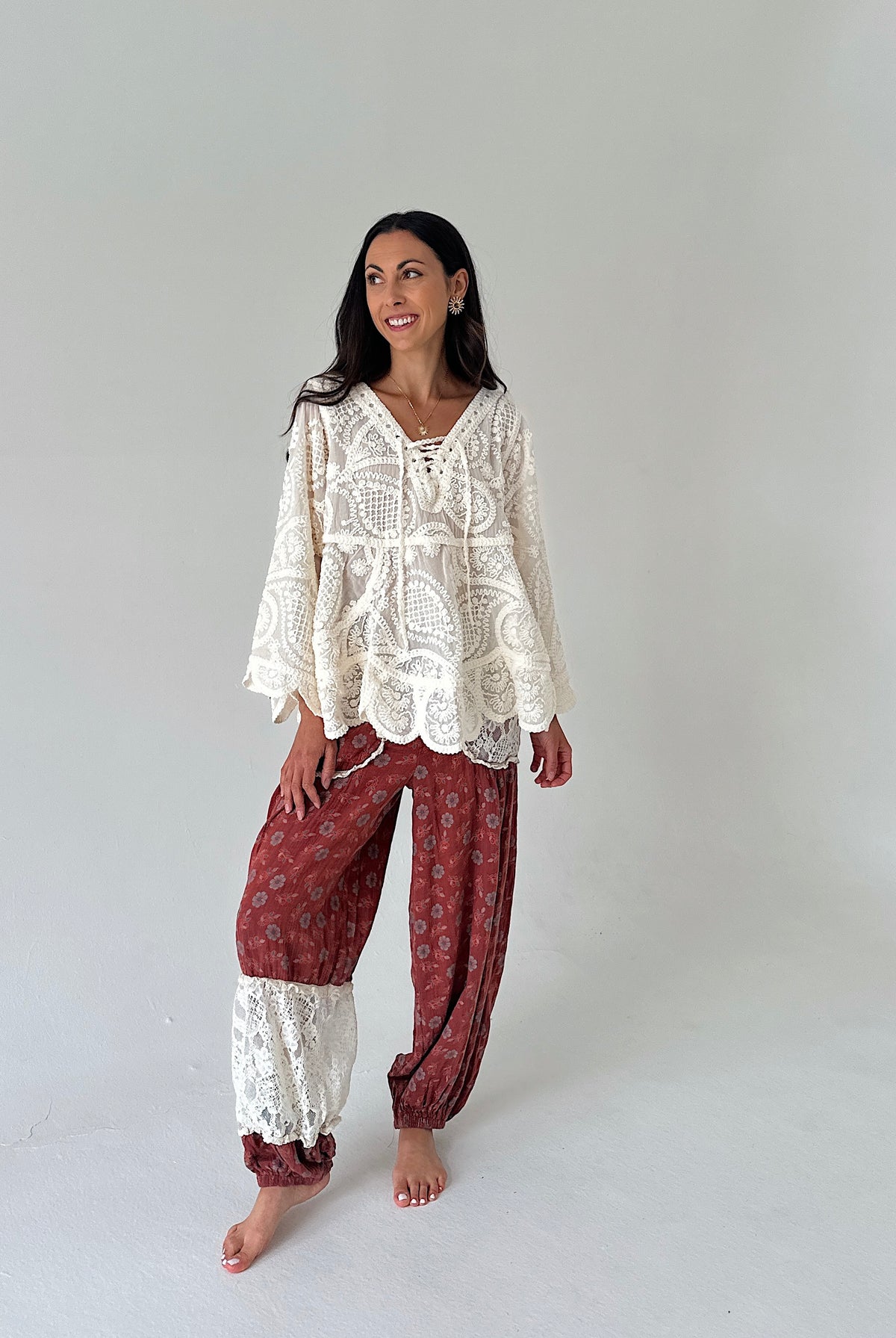 A woman modeling a bohemian-style two-piece outfit consisting of a cream lace long-sleeve top and rust-red patterned harem pants with lace cuffs, standing against a plain light gray studio background.