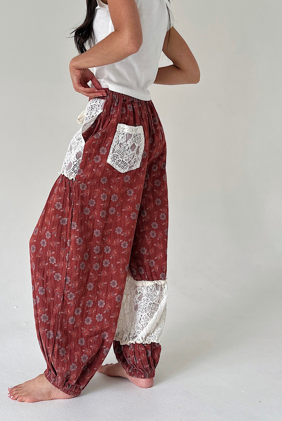 A person wearing loose, high-waisted harem-style pants in a rust red floral print with white lace pocket and lace lower-leg panels, paired with a white sleeveless top against a plain light gray studio background.