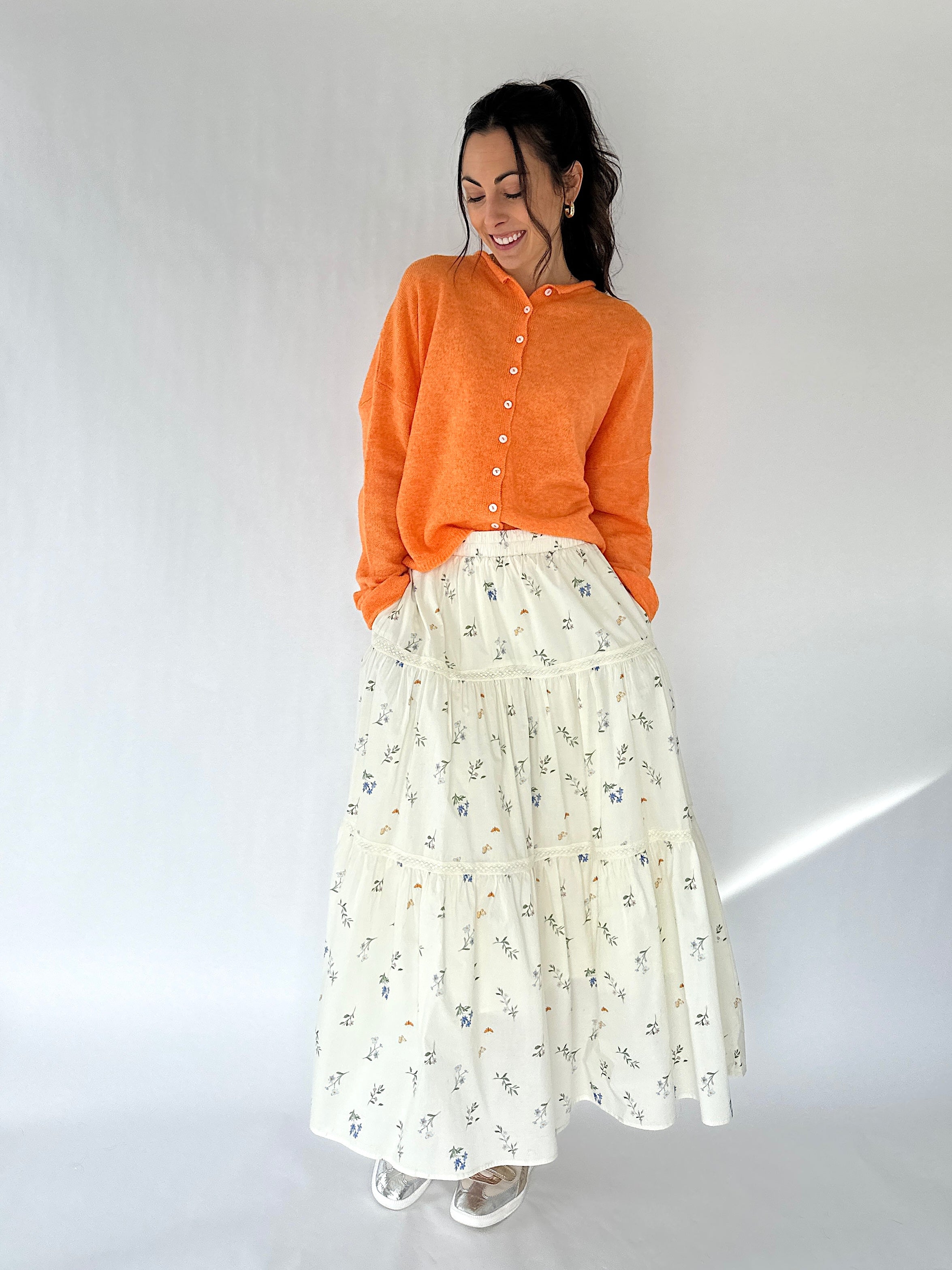 Woman wearing an orange sweater and white floral skirt on a plain background