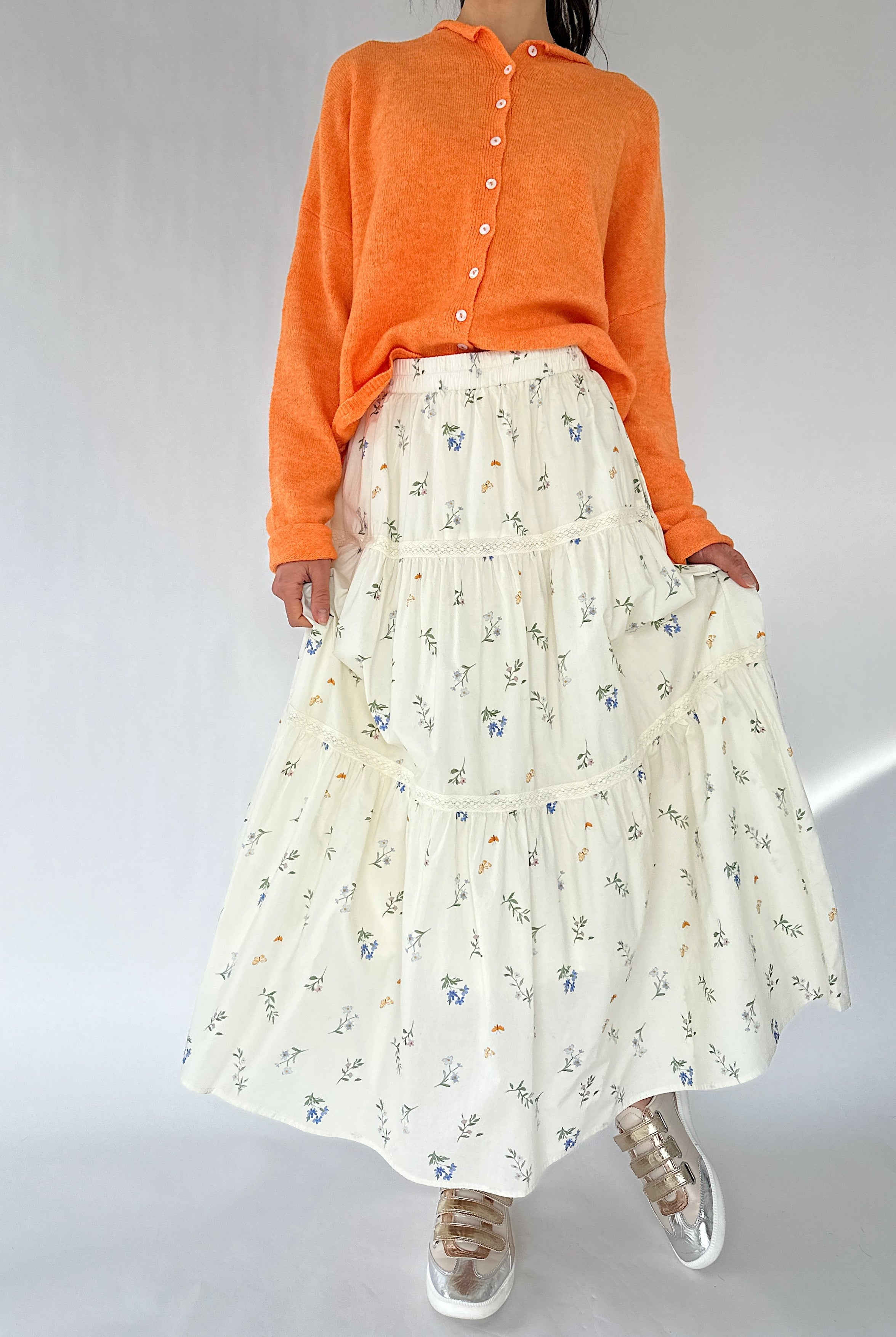 Person wearing an orange sweater and a white floral skirt on a plain background