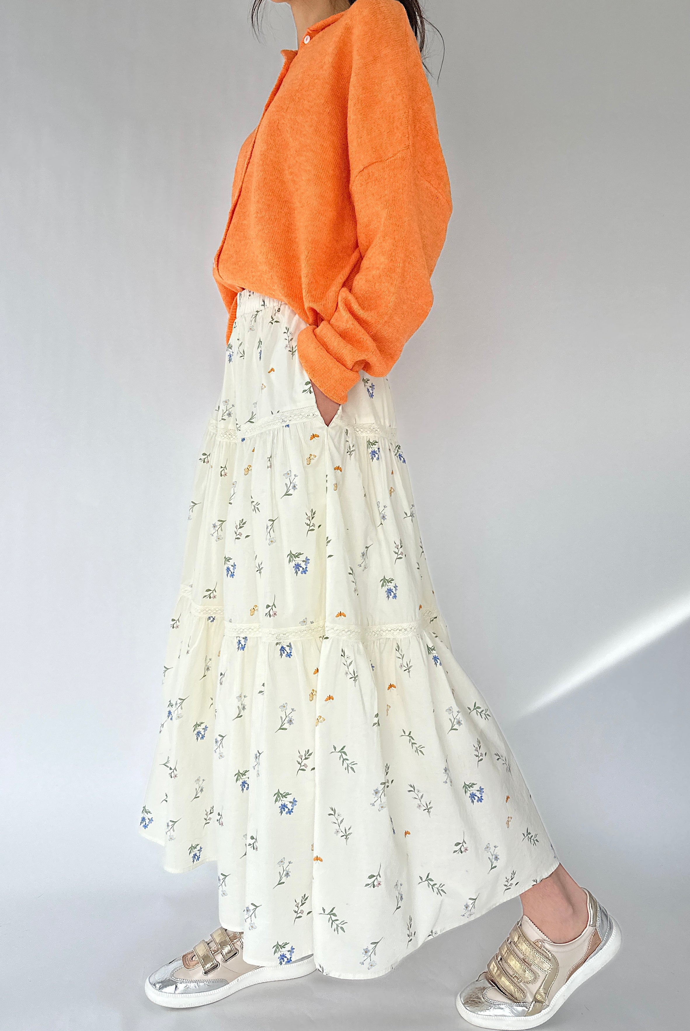 Person wearing an orange sweater and floral skirt on a plain background showing side pockets