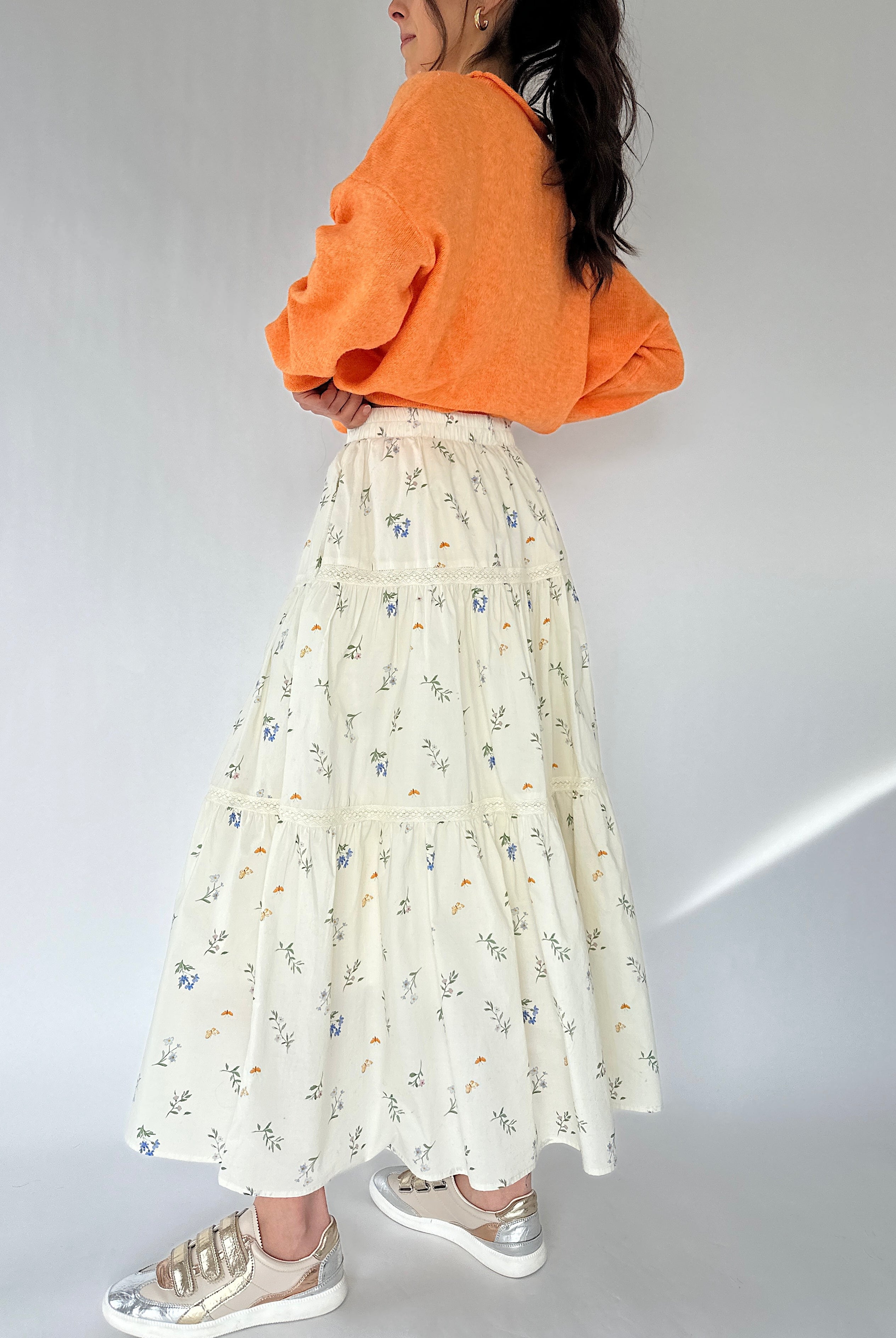 Person wearing an orange sweatshirt and a long floral skirt on a plain background