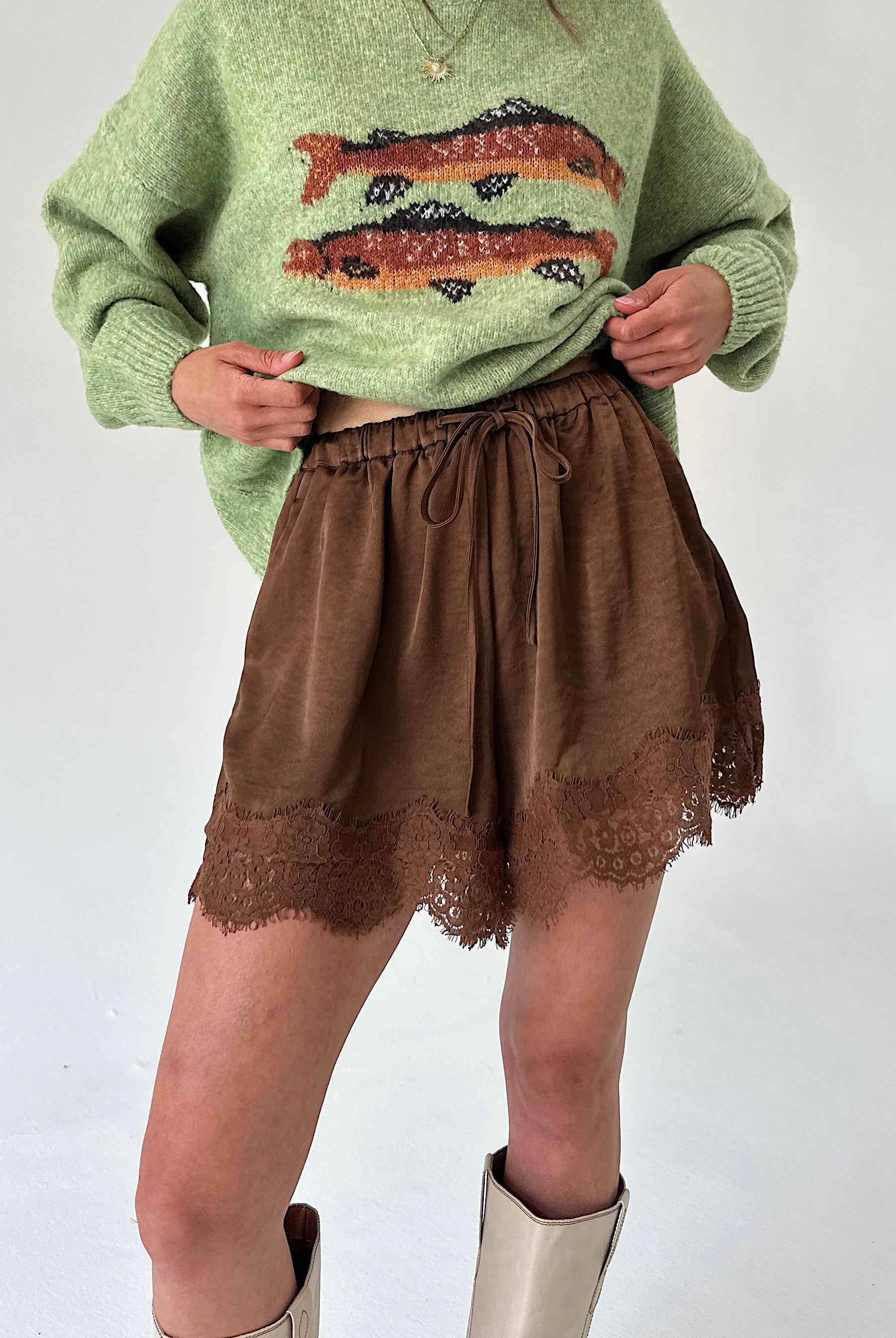 A person wearing a light green knit sweater with a fish pattern and brown shorts with a lace hem, paired with beige boots, against a plain light background.