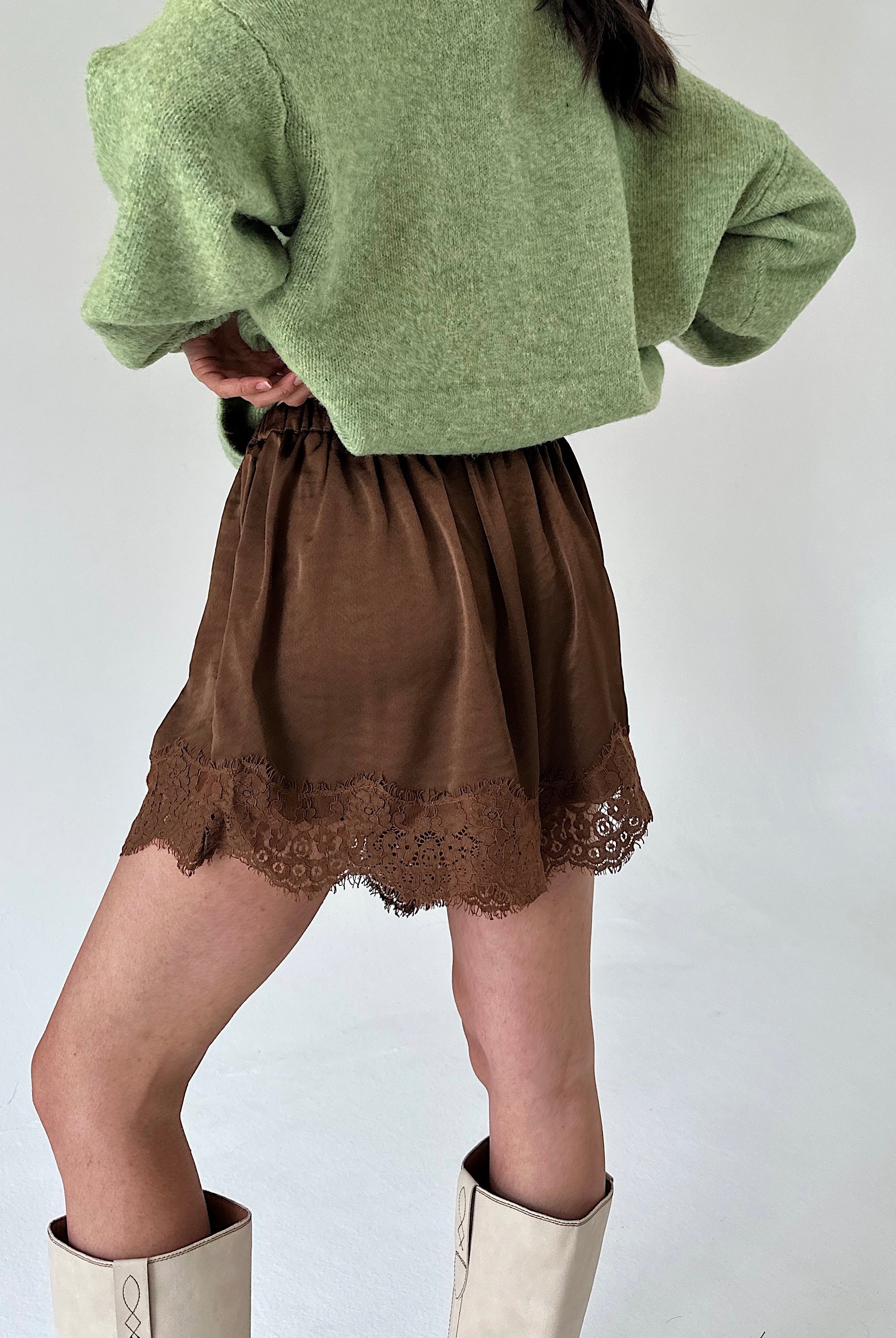 A person shown from the back wearing a light green fuzzy cropped sweater, a brown skirt with a lace hem, and off-white knee-high boots against a plain light gray background.