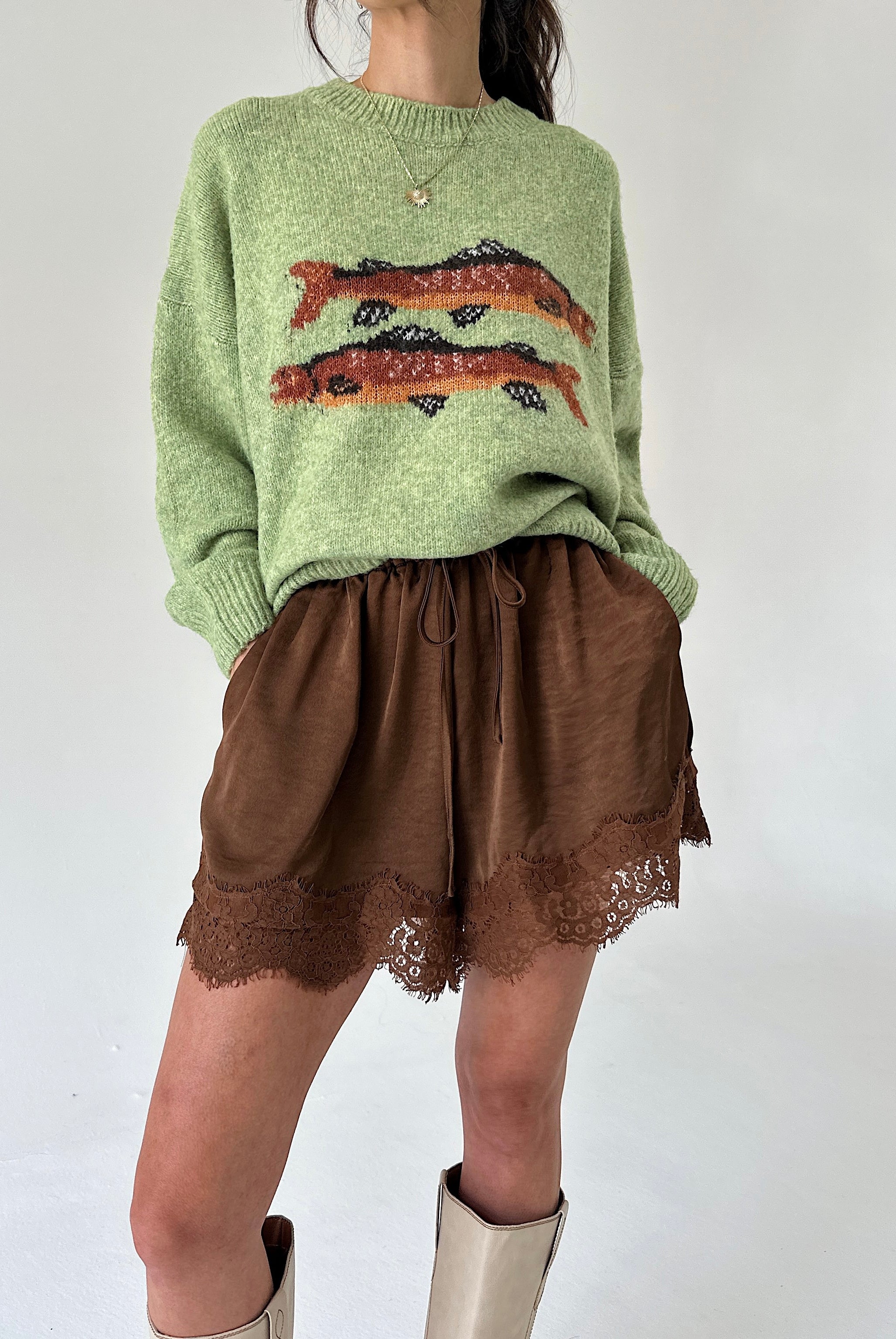 A model wearing a light green knit sweater with a brown and orange fish pattern on the chest, paired with brown shorts featuring a lace hem and beige boots, set against a plain white background.