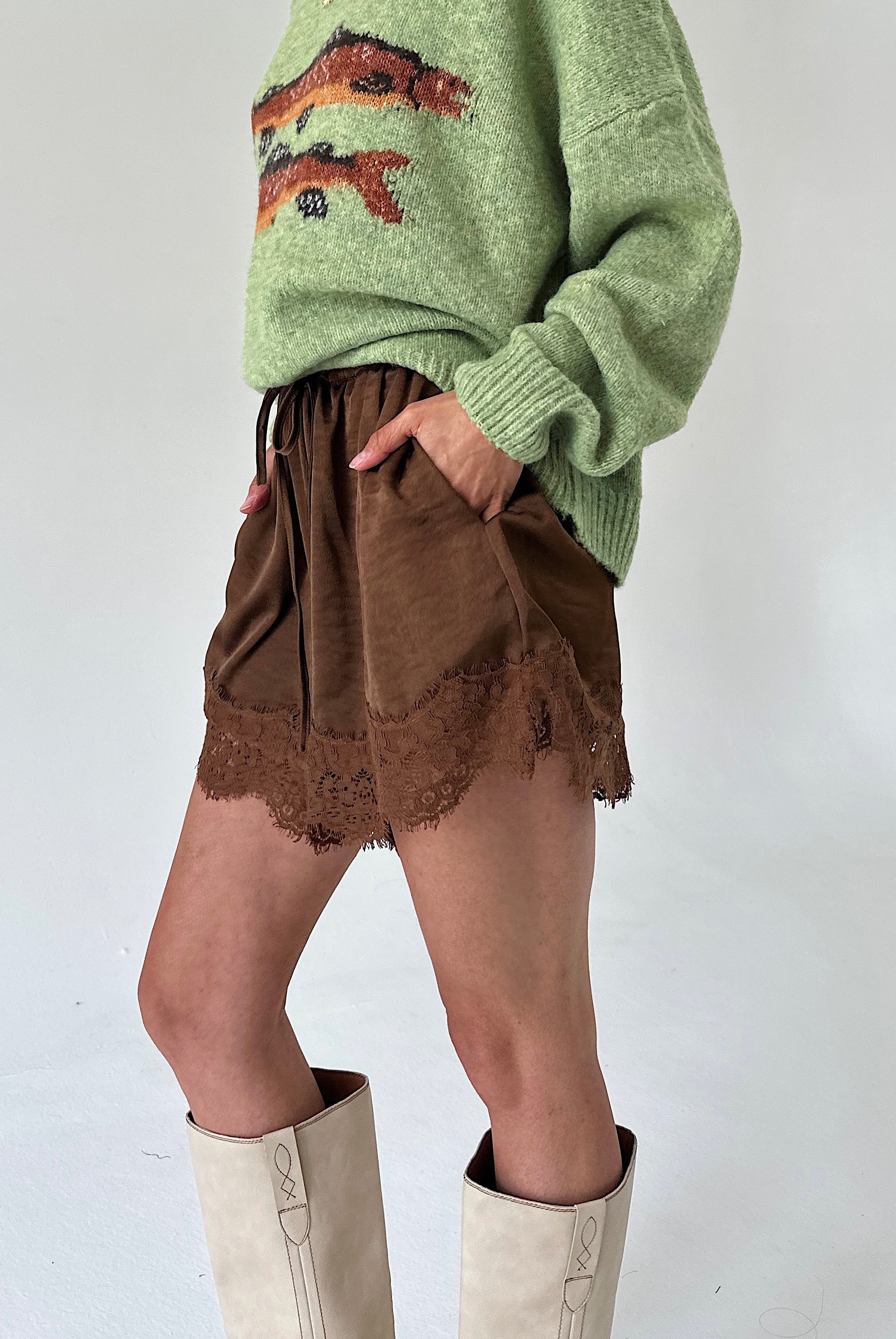A person wearing brown satinshorts with a lace hem, paired with a light green sweater and off-white knee-high boots, shown against a plain light background.
