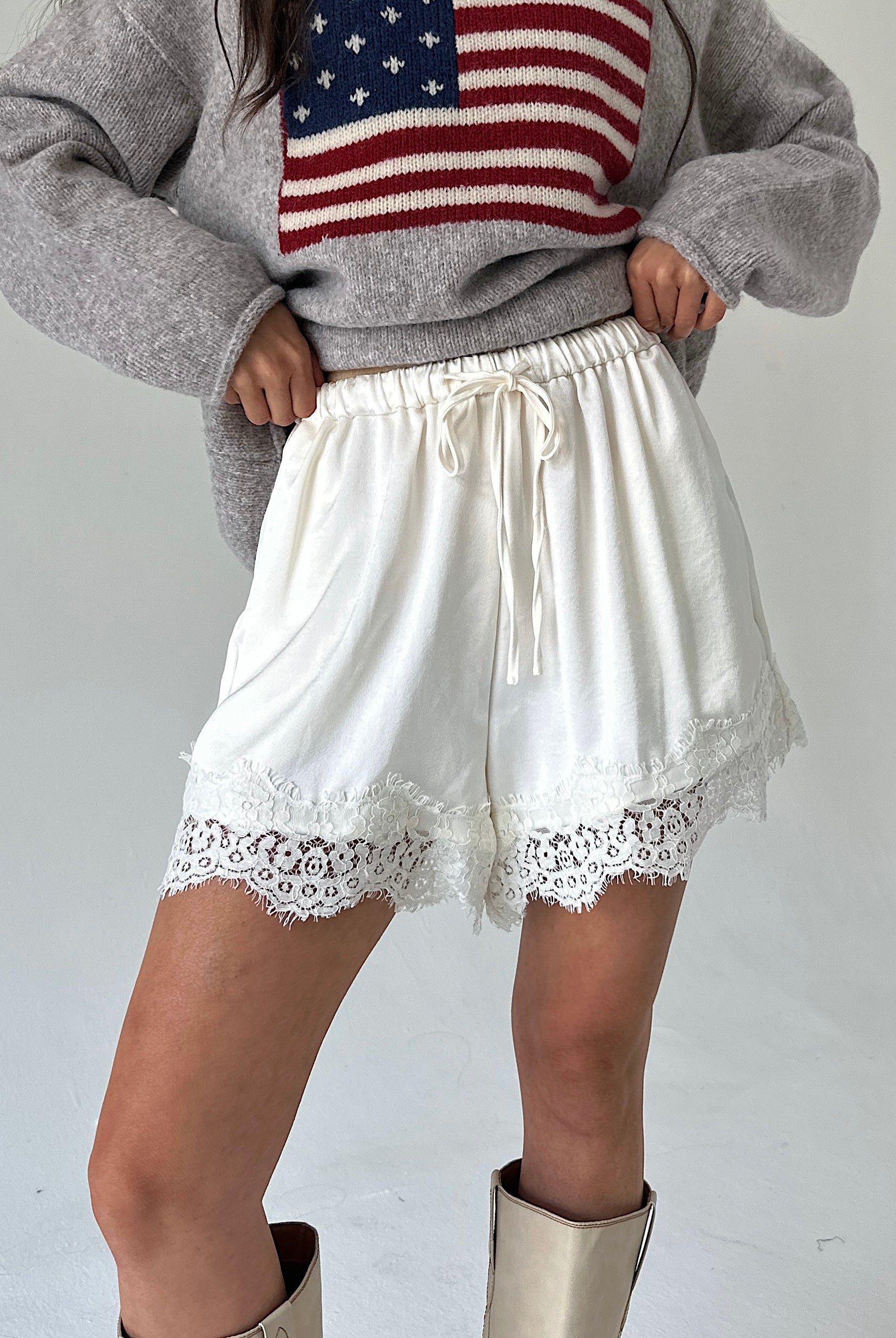 A person wearing white drawstring shorts with lace trim, paired with a gray sweater featuring an American flag graphic and beige boots, photographed against a plain light background.