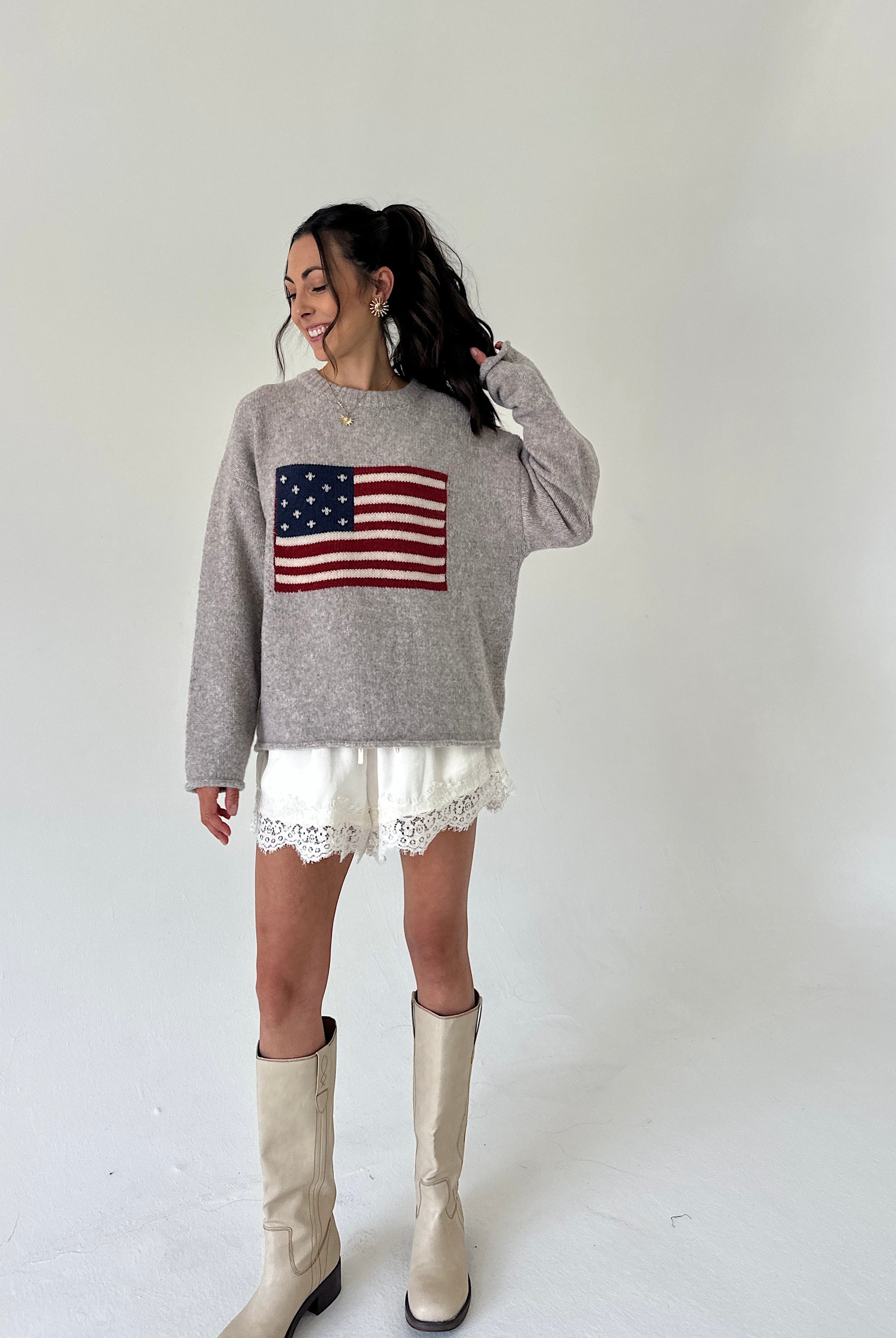 A woman modeling a heather gray crewneck sweater featuring a red, white, and blue American flag graphic on the front, paired with white lace-trimmed shorts and beige knee-high boots, set against a plain white studio background.