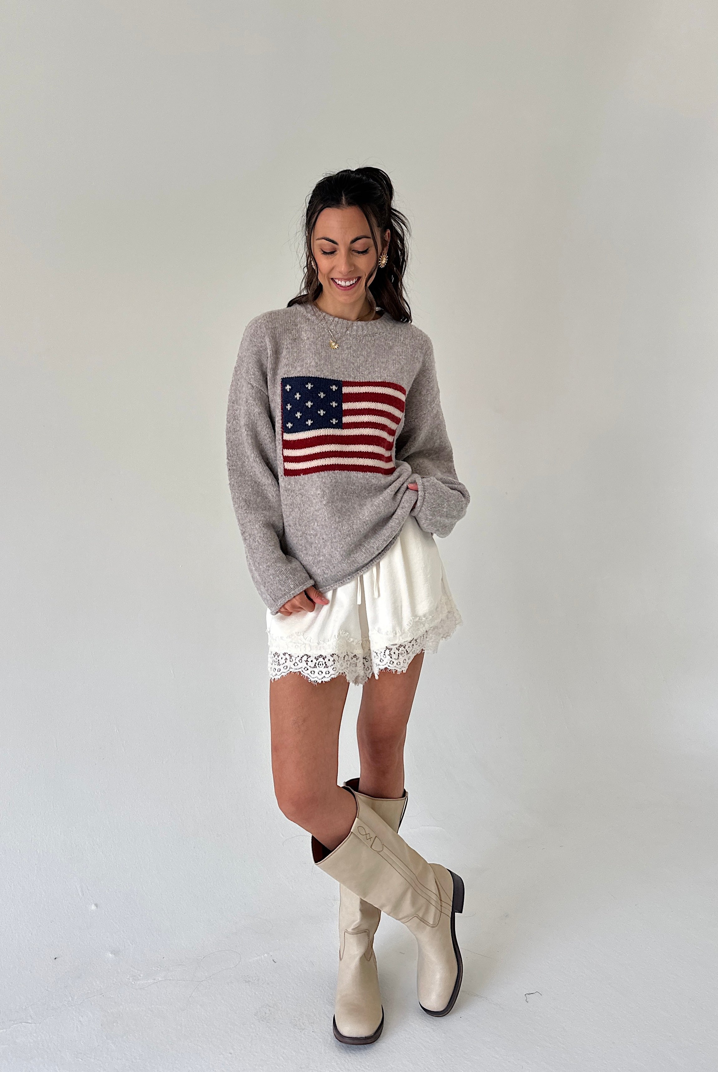A woman modeling a light gray knit sweater with a red, white, and blue American flag graphic on the front, paired with white lace-trimmed shorts and beige knee-high boots, against a plain light gray studio background.
