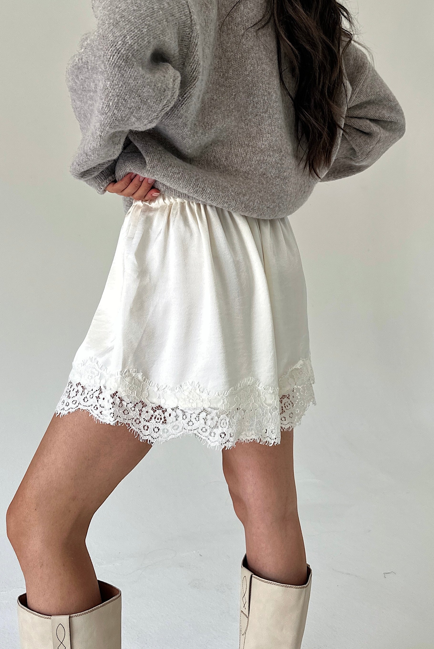 A person wearing white satin shorts with a lace hem, styled with a gray knit sweater and beige boots against a plain light background.