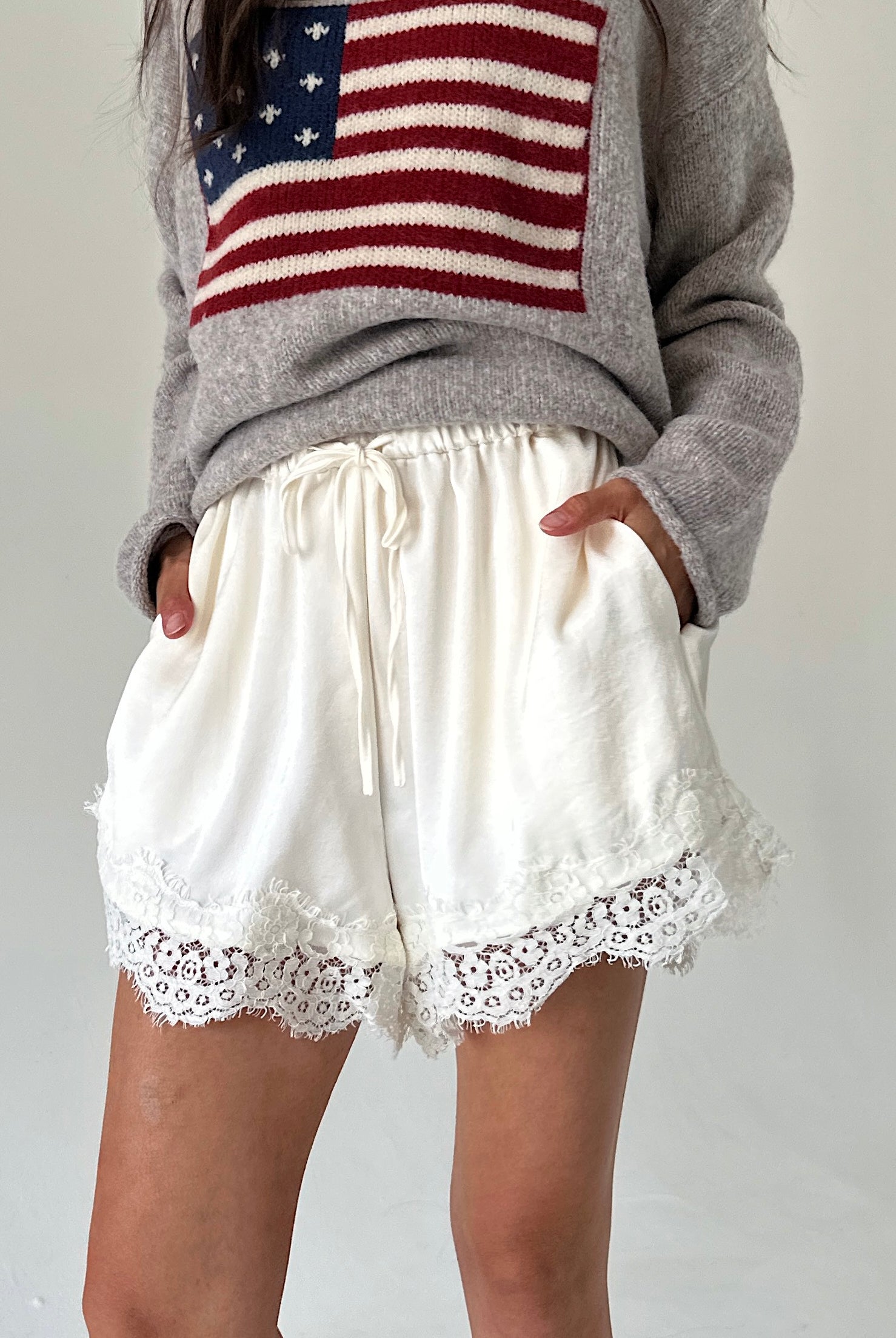 A person wearing white drawstring shorts with lace trim and a gray sweater featuring an American flag graphic, standing against a plain light background.
