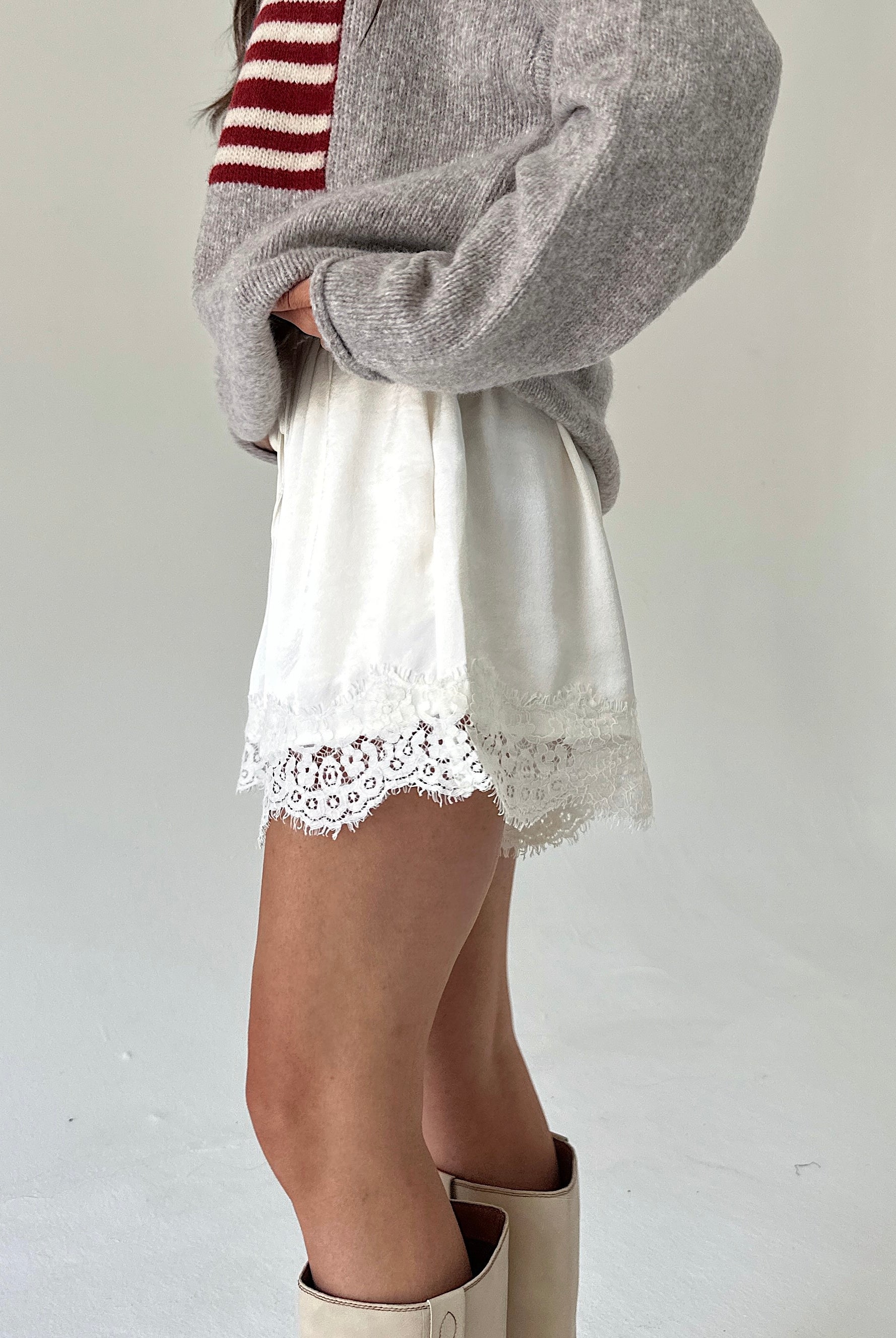 A person wearing a taupe knit sweater with a red and white stripe, white shorts with a lace hem, and beige boots, shown from the chest down against a plain light background.