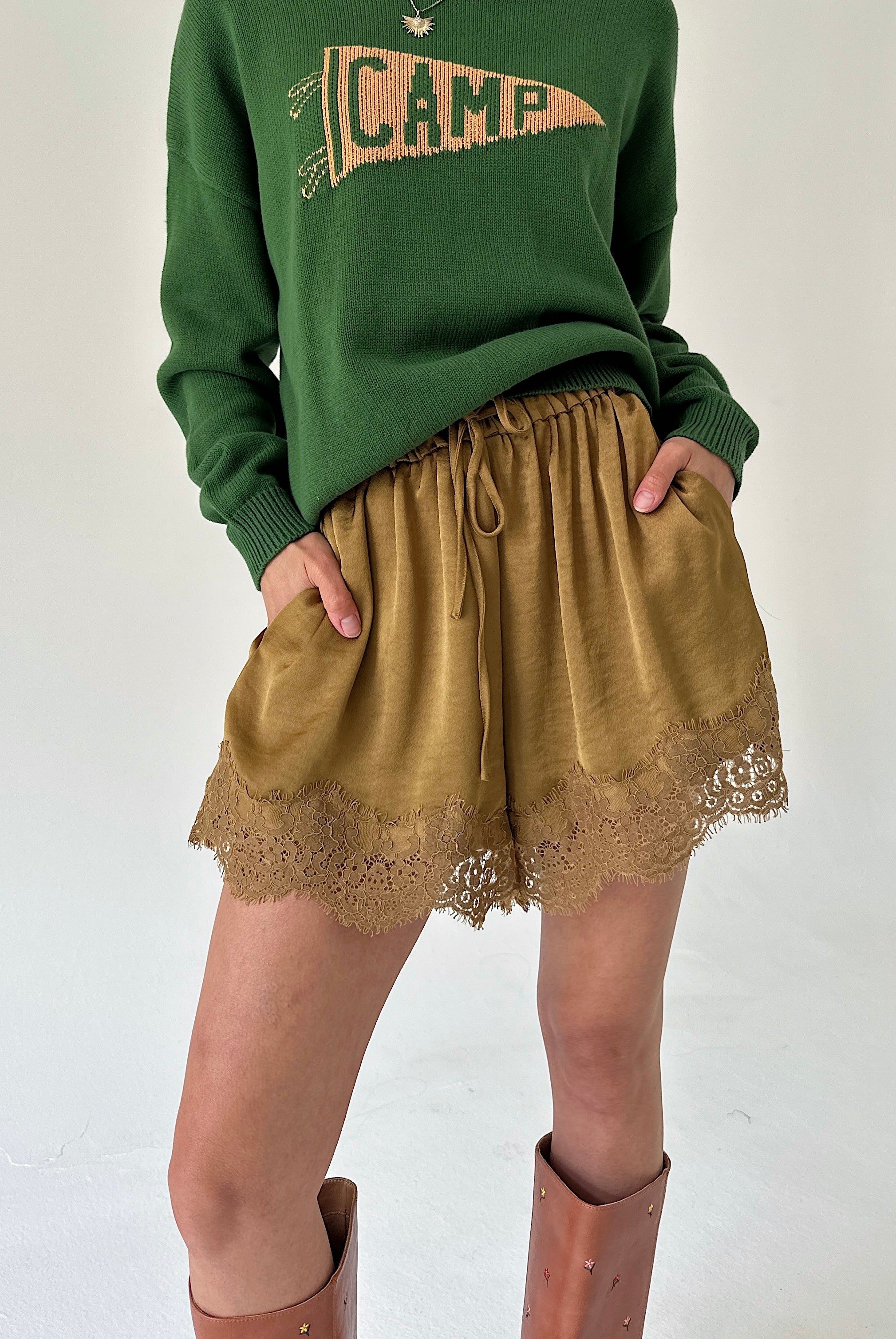 A person wearing a green crewneck sweater with a 'CAMP' pennant graphic and mustard satin shorts with a lace hem, paired with brown knee-high boots, against a plain off-white background.