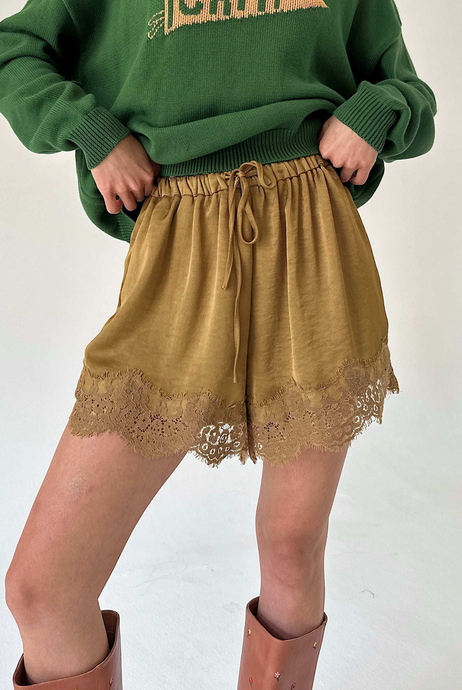 A person wearing olive green knit sweater and mustard drawstring shorts with lace trim, paired with brown boots, against a plain light background.
